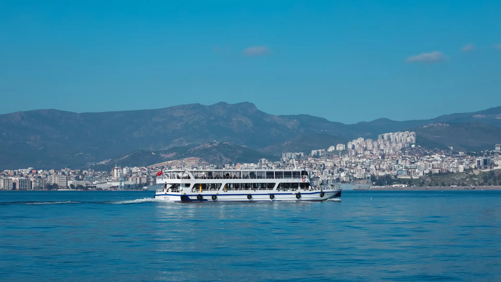Modern passenger ferry approaching sunny İzmir waterfront across blue bay water