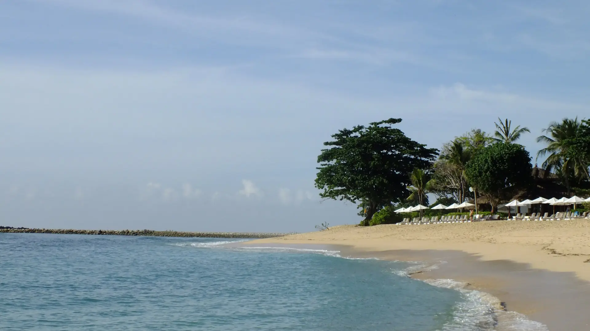 A curved pale-sand beach, palms and beach umbrellas beside blue water on the wider Nusa Dua coast in Bali
