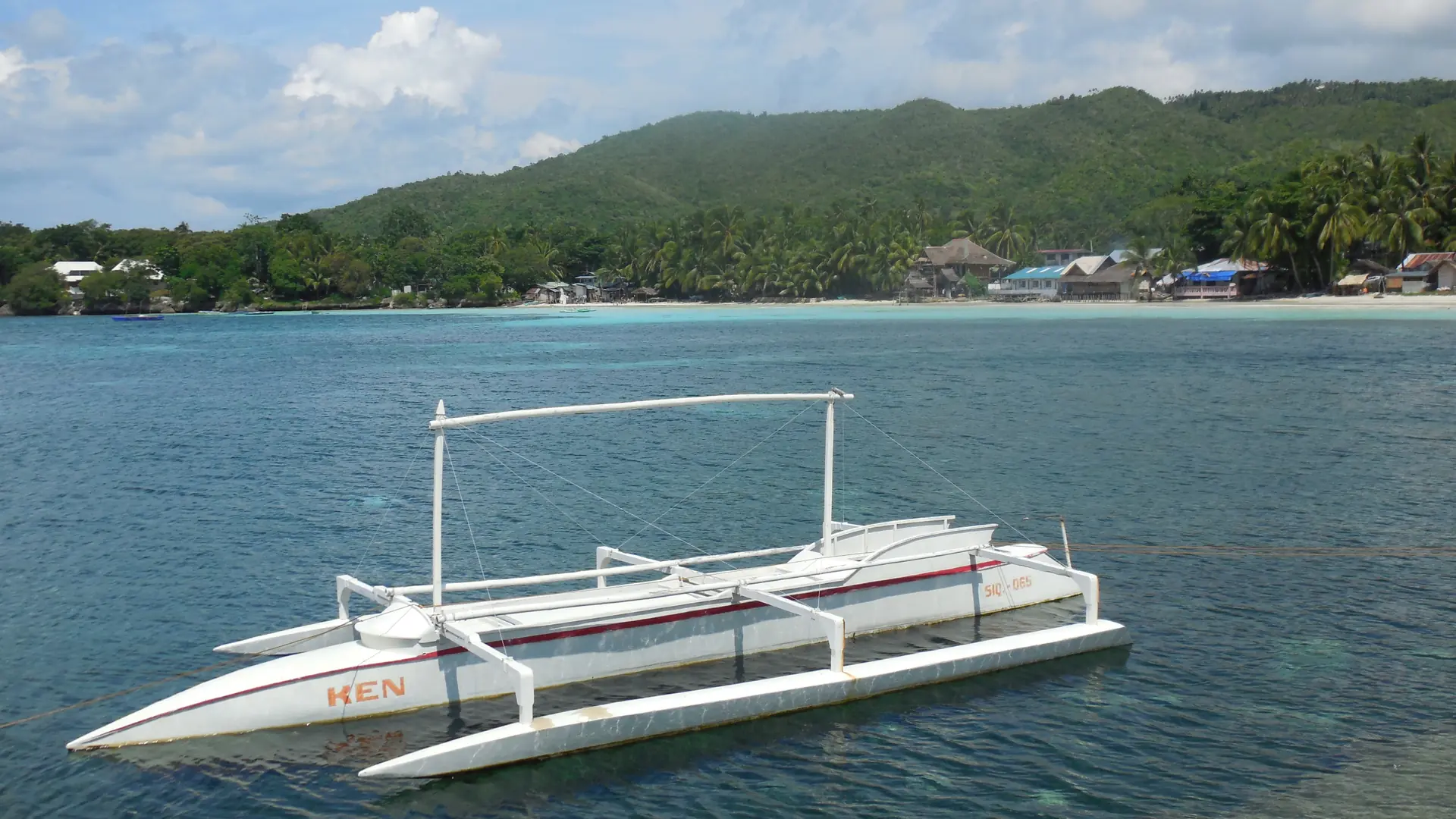 A white outrigger boat on blue water below Siquijor’s green hills and coastal buildings