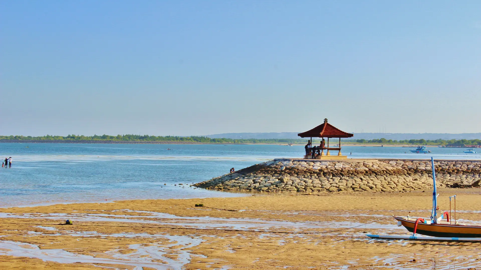 A red-roofed pavilion and traditional boat beside exposed sand and shallow blue water on the wider Sanur coast in Bali
