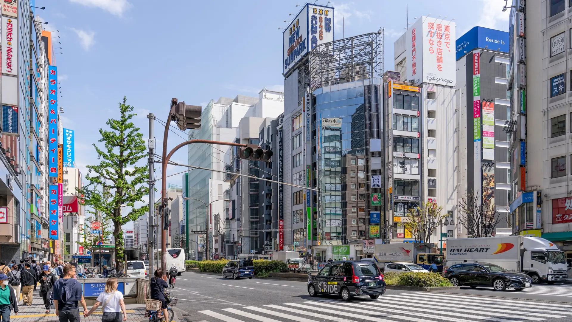 Sunlit Shinjuku avenue beside Sunpark buildings, with shops, pedestrians and traffic in Tokyo