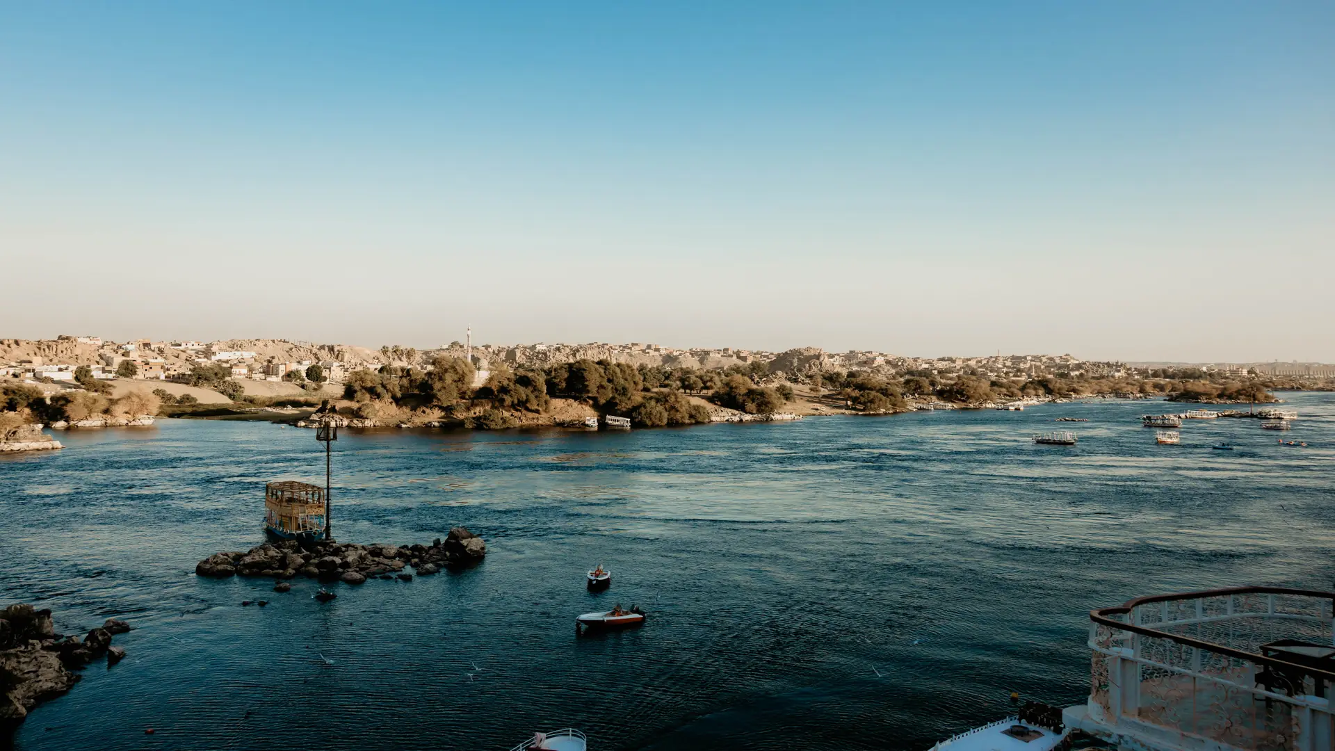 Boats on the Nile beside the green Aswan shoreline beneath a bright blue sky.