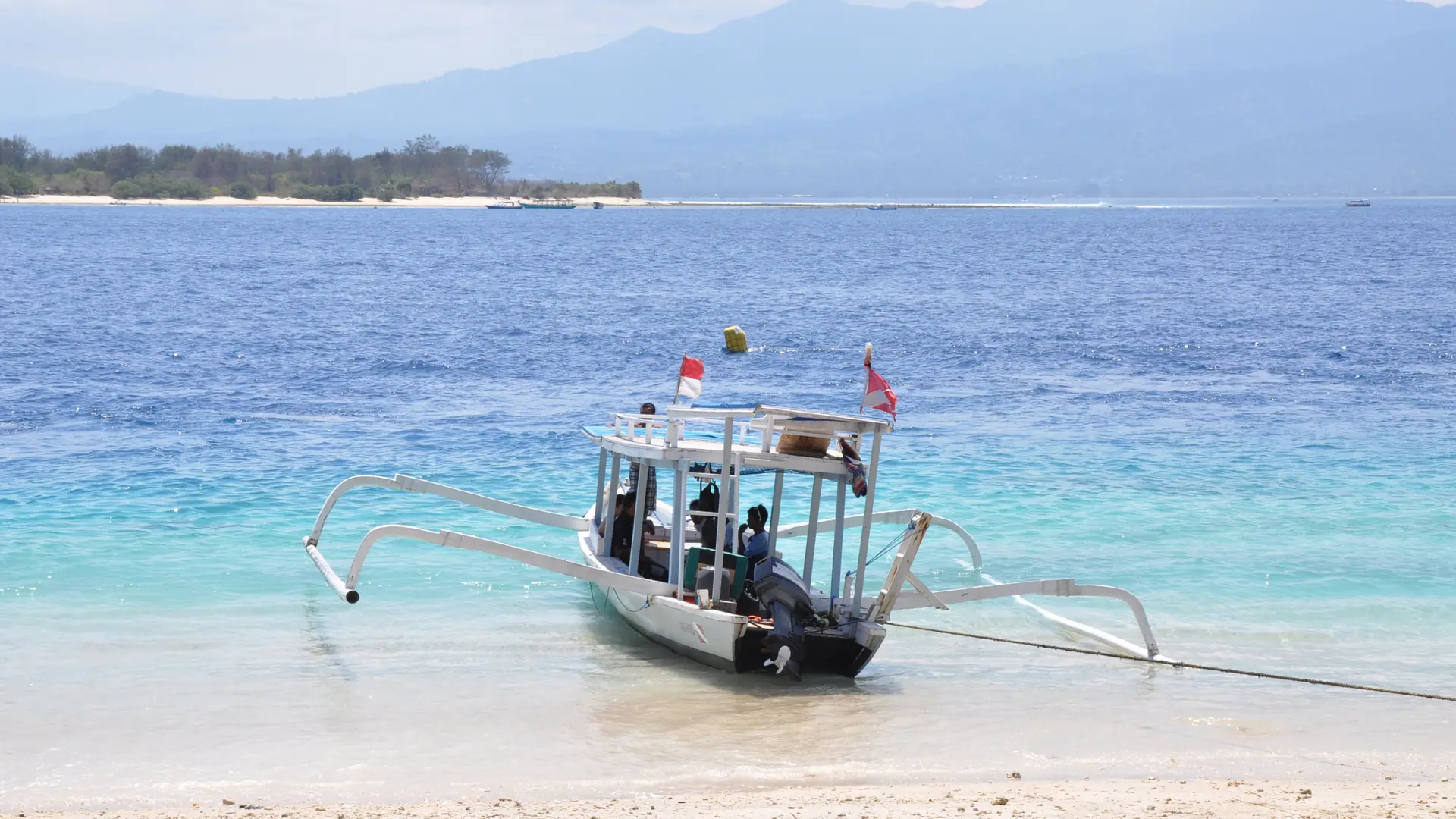 White outrigger boat with Indonesian flags beside a Gili Trawangan beach and Lombok mountains beyond