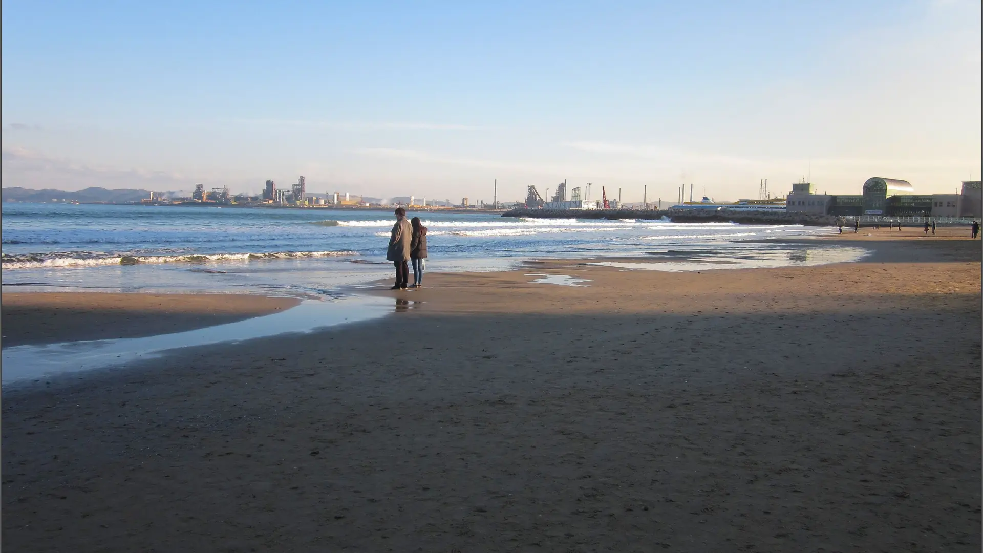 People walking on Yeongildae Beach in daylight, with the industrial shoreline across Pohang bay