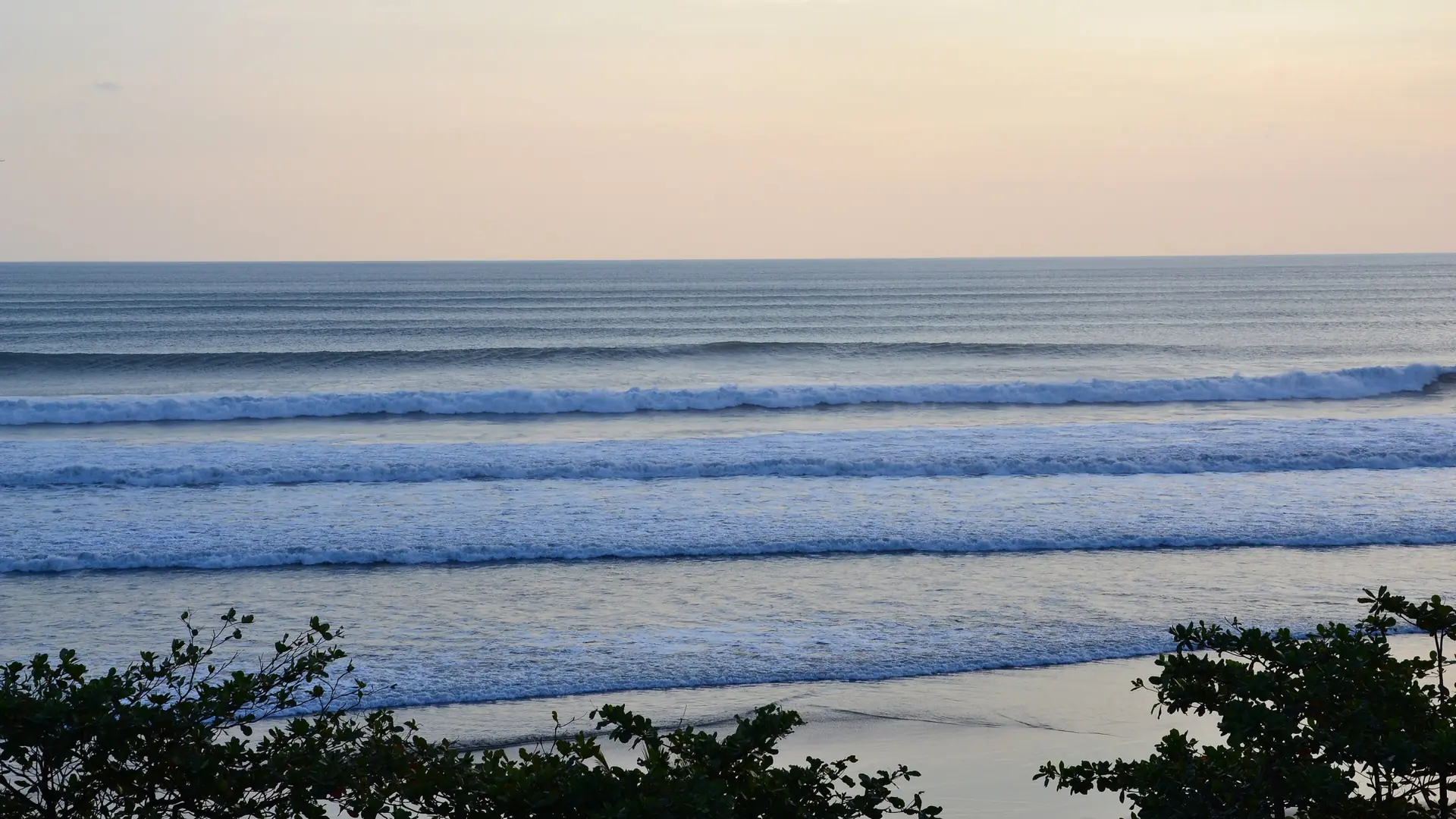 Lines of surf along Seminyak Beach beneath a pale golden sky, viewed from Anantara Seminyak