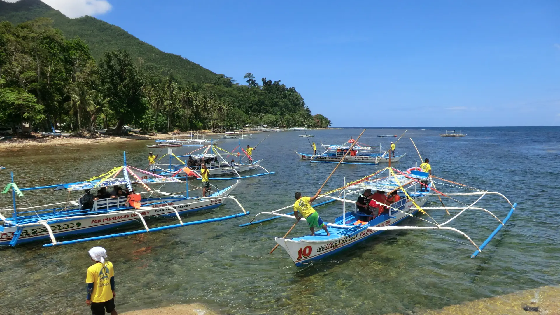 Passenger outrigger boats and their crews in the sunlit shallows of Sabang, with a forested coastal hill behind