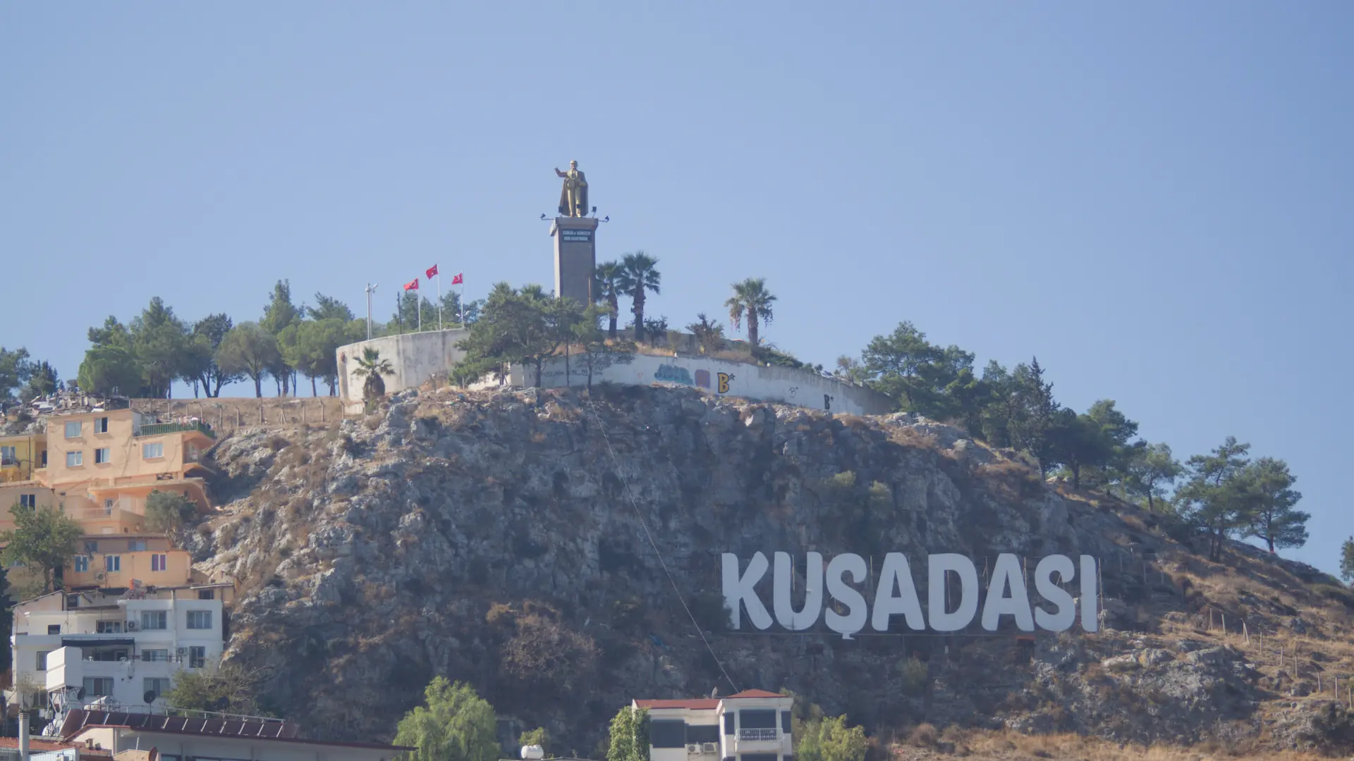Sunlit Kuşadası hillside lettering and Atatürk statue, shown as a wider city scene