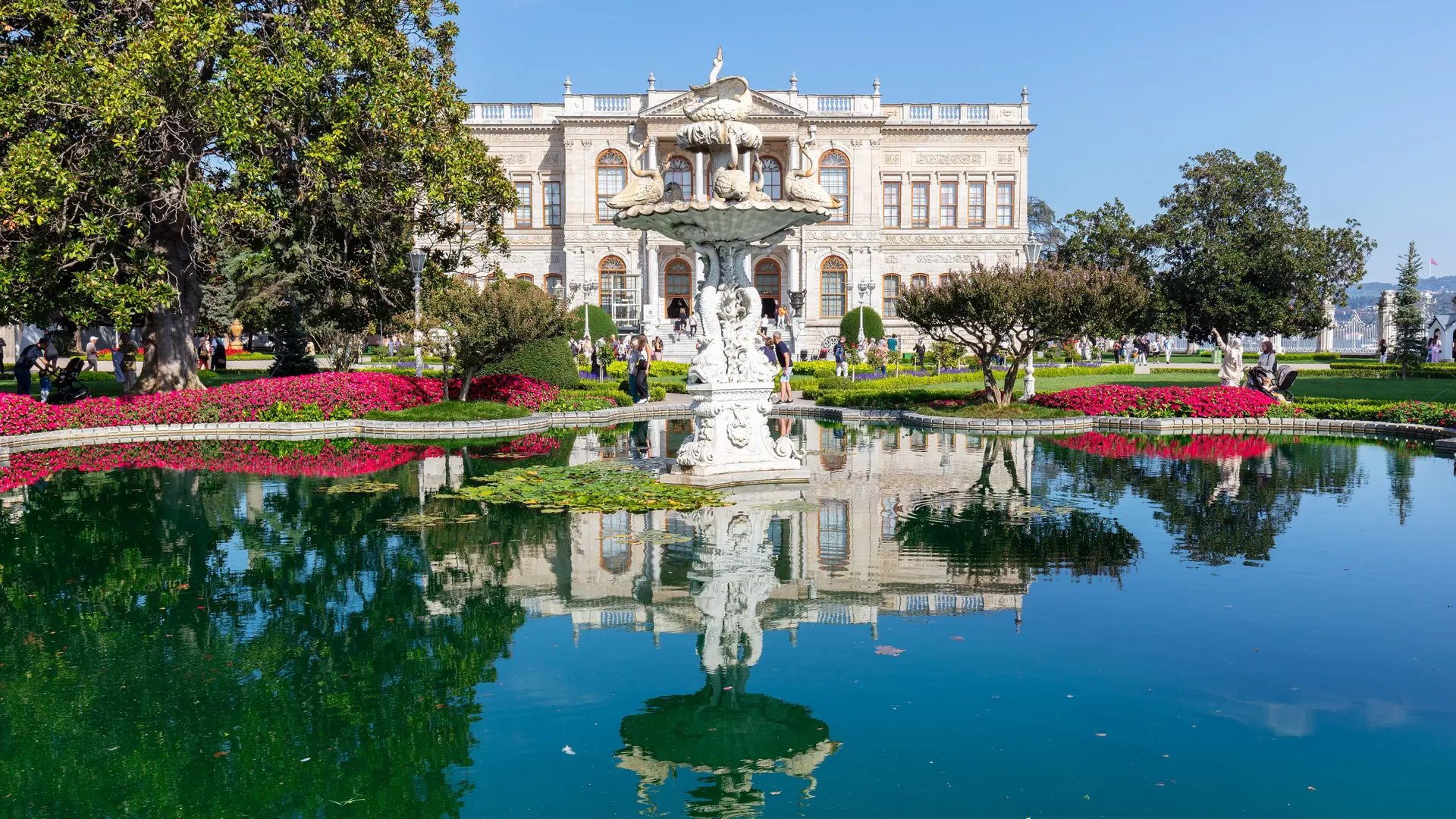 Dolmabahçe Palace and its garden fountain reflected in a pond under blue skies