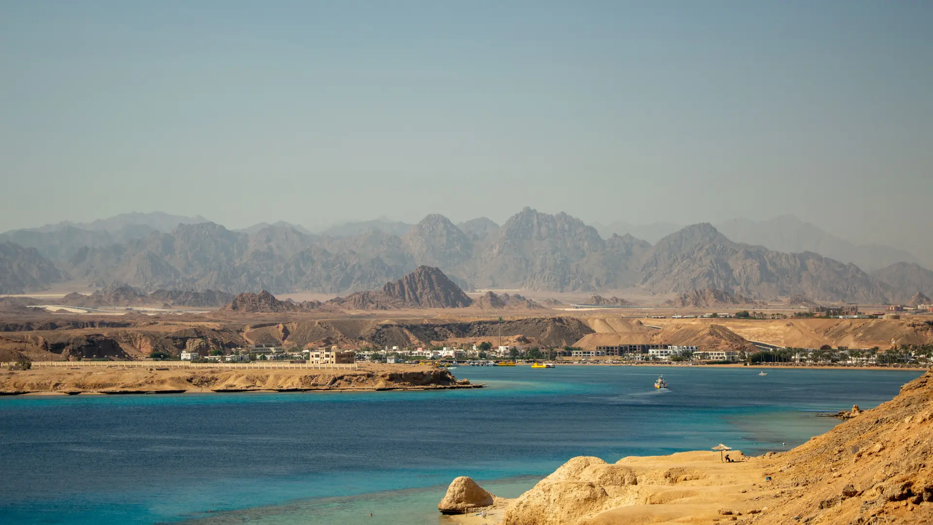 Sunlit Sharm el-Sheikh coastal bay with turquoise water, rocky shoreline and Sinai mountains beyond.