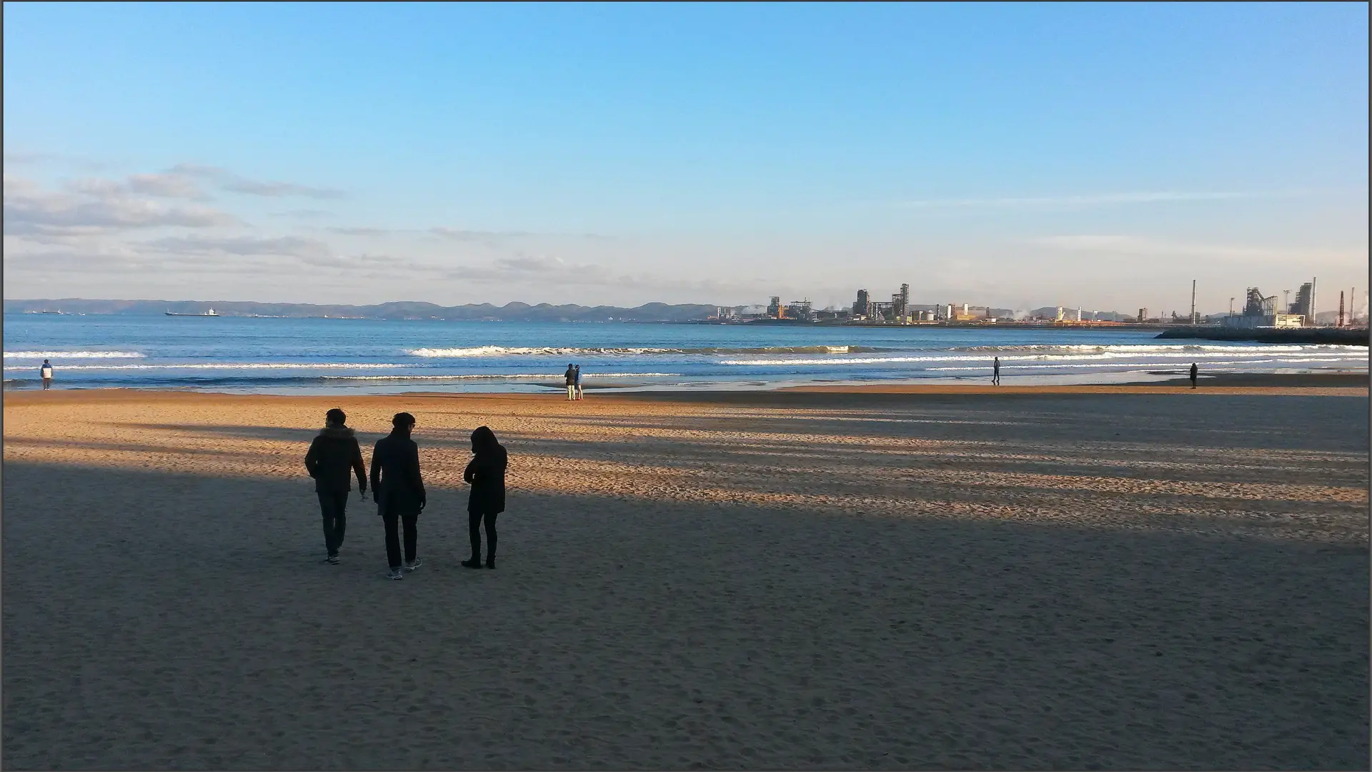 Three people on the sand at Yeongildae Beach, with water and the industrial shoreline beyond