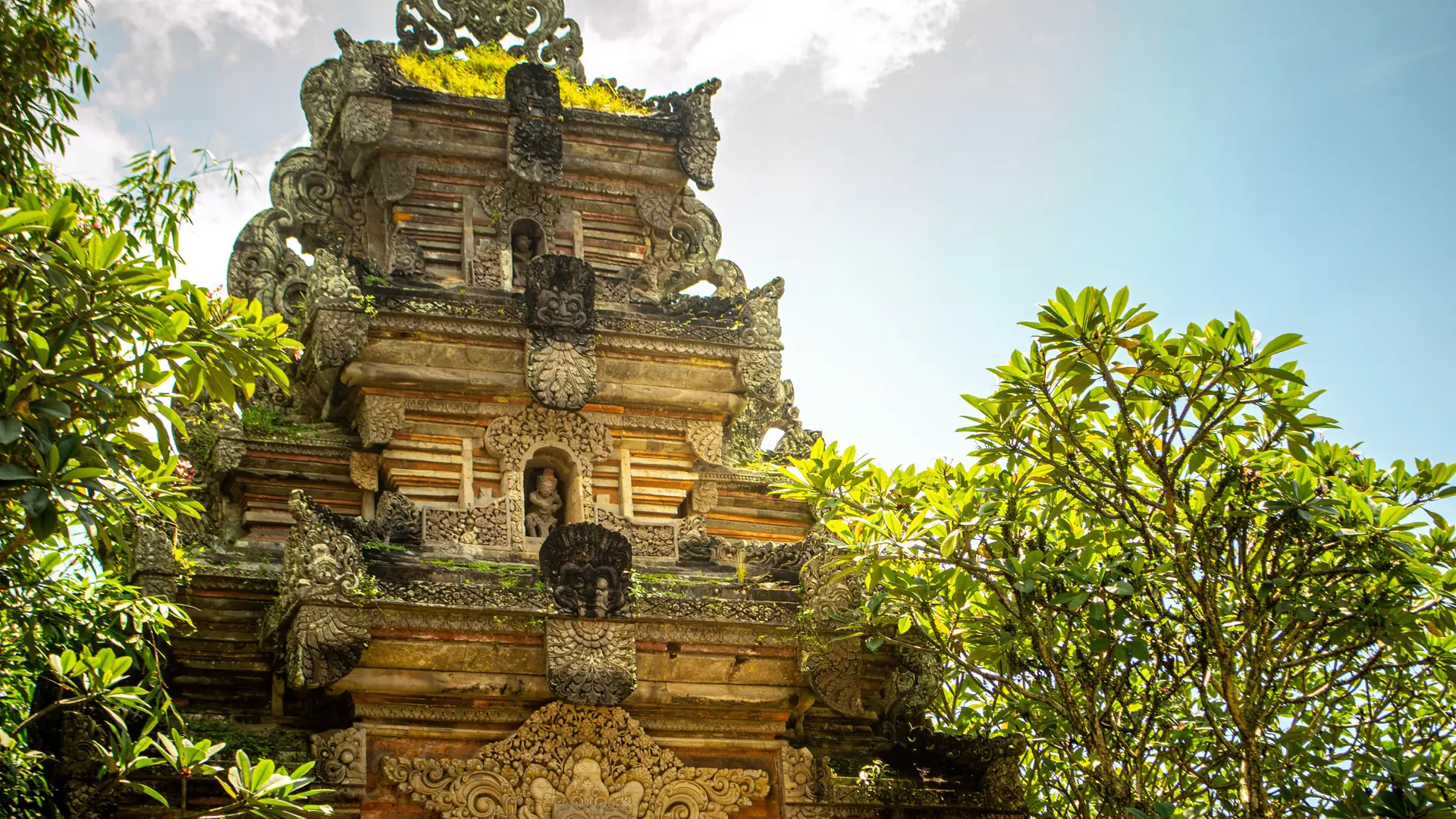Sunlit carved stone gateway and guardian at Ubud Palace among frangipani trees