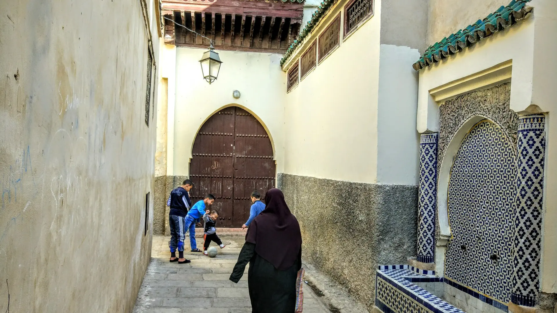 Residents on a lane beside a tiled street fountain in Fez medina