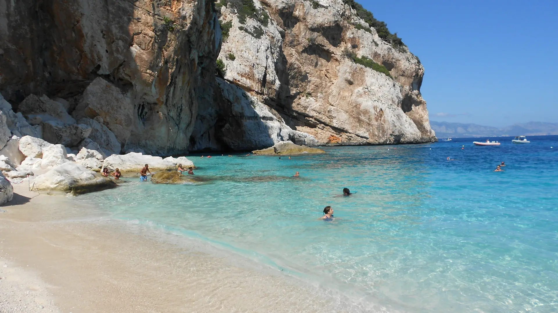 Swimmers in clear turquoise water beside pale rocks and limestone cliffs at Cala Mariolu, Sardinia.