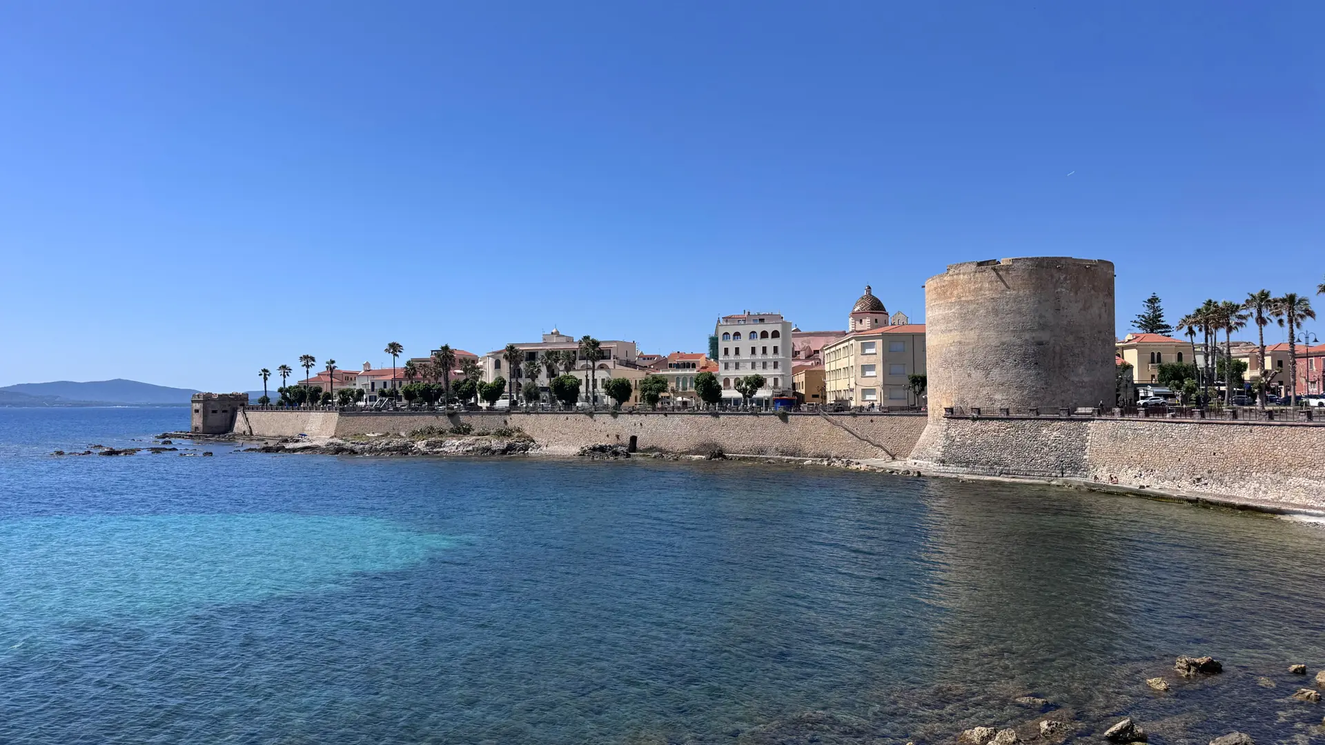 Torre Sulis and Alghero’s stone waterfront beside clear blue water on a sunny day.