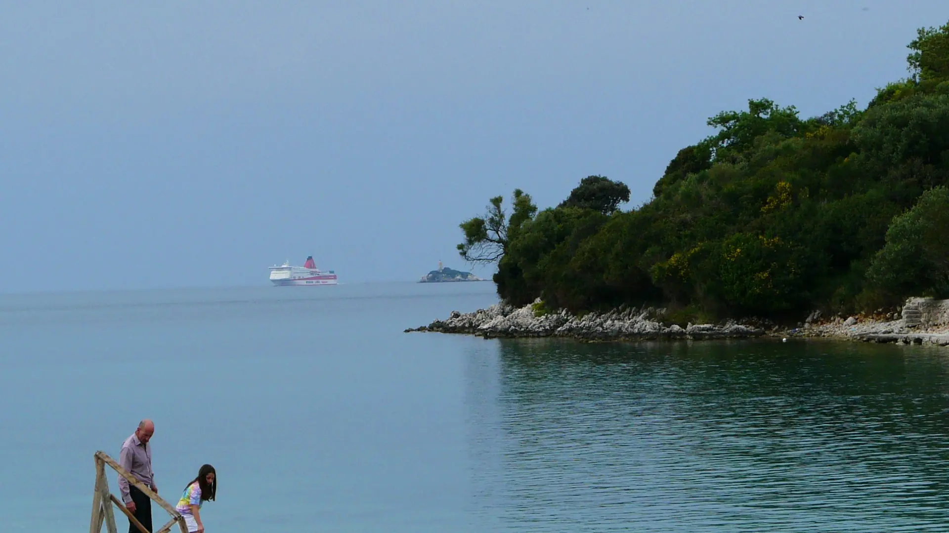 Swimmers beside a rocky Ksamil shore, with a wooded islet and a ferry visible in the Corfu Strait