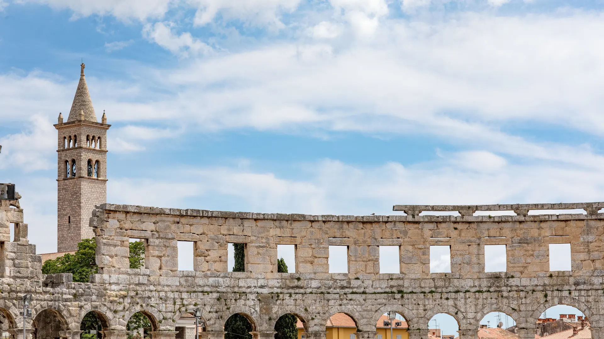 Pula Arena’s outer stone arches and a nearby bell tower under a blue sky