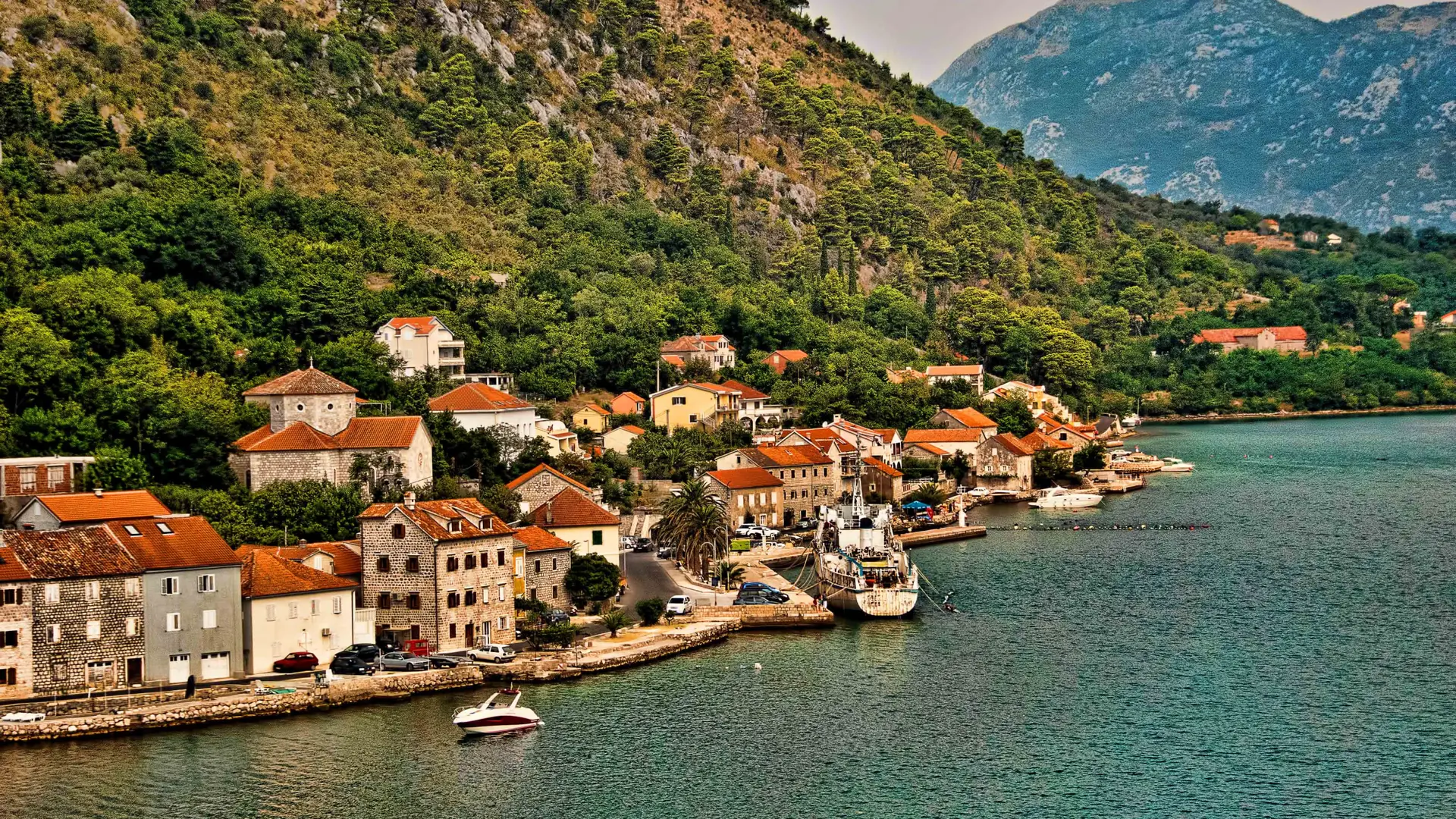 Stone houses and small jetties along the Dobrota waterfront, with calm bay water and mountains behind