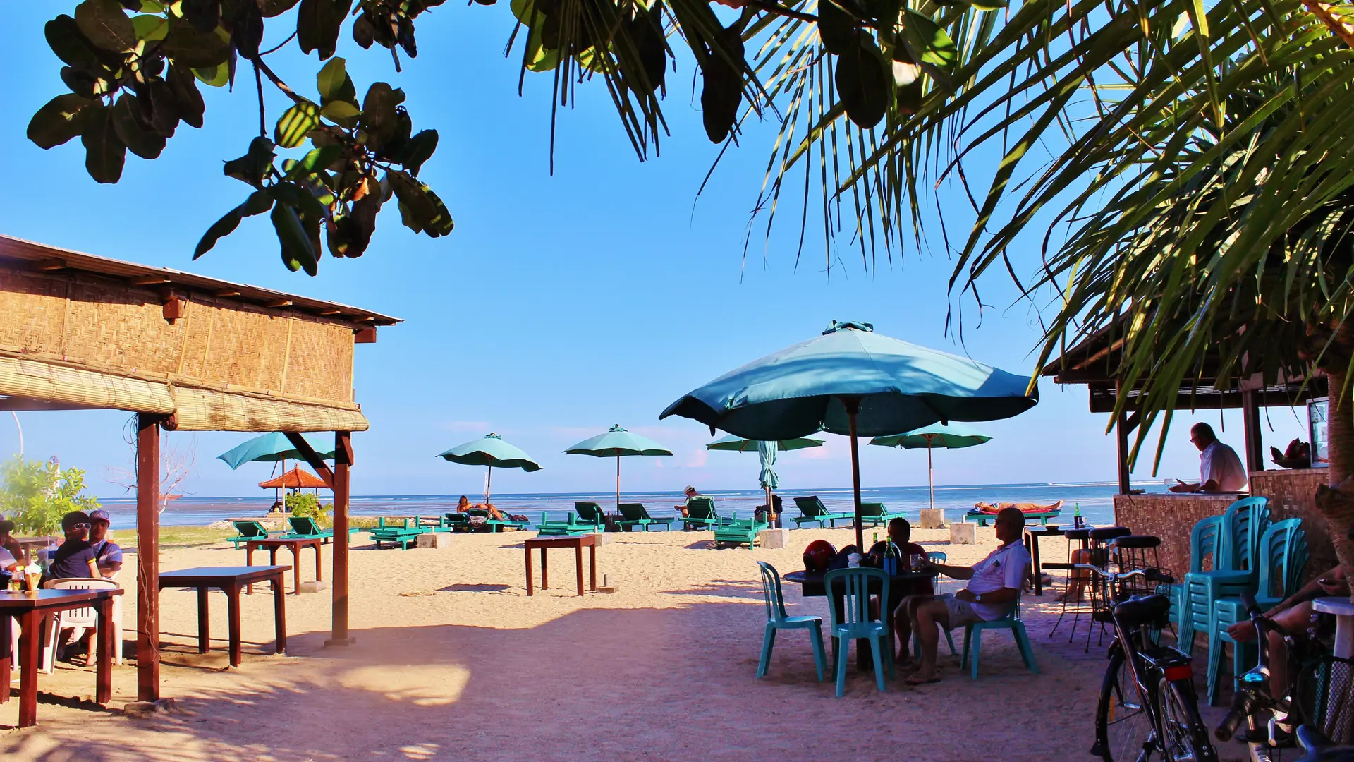 Beachside cafe seating, green umbrellas and palms overlooking the sunny wider Sanur shoreline in Bali