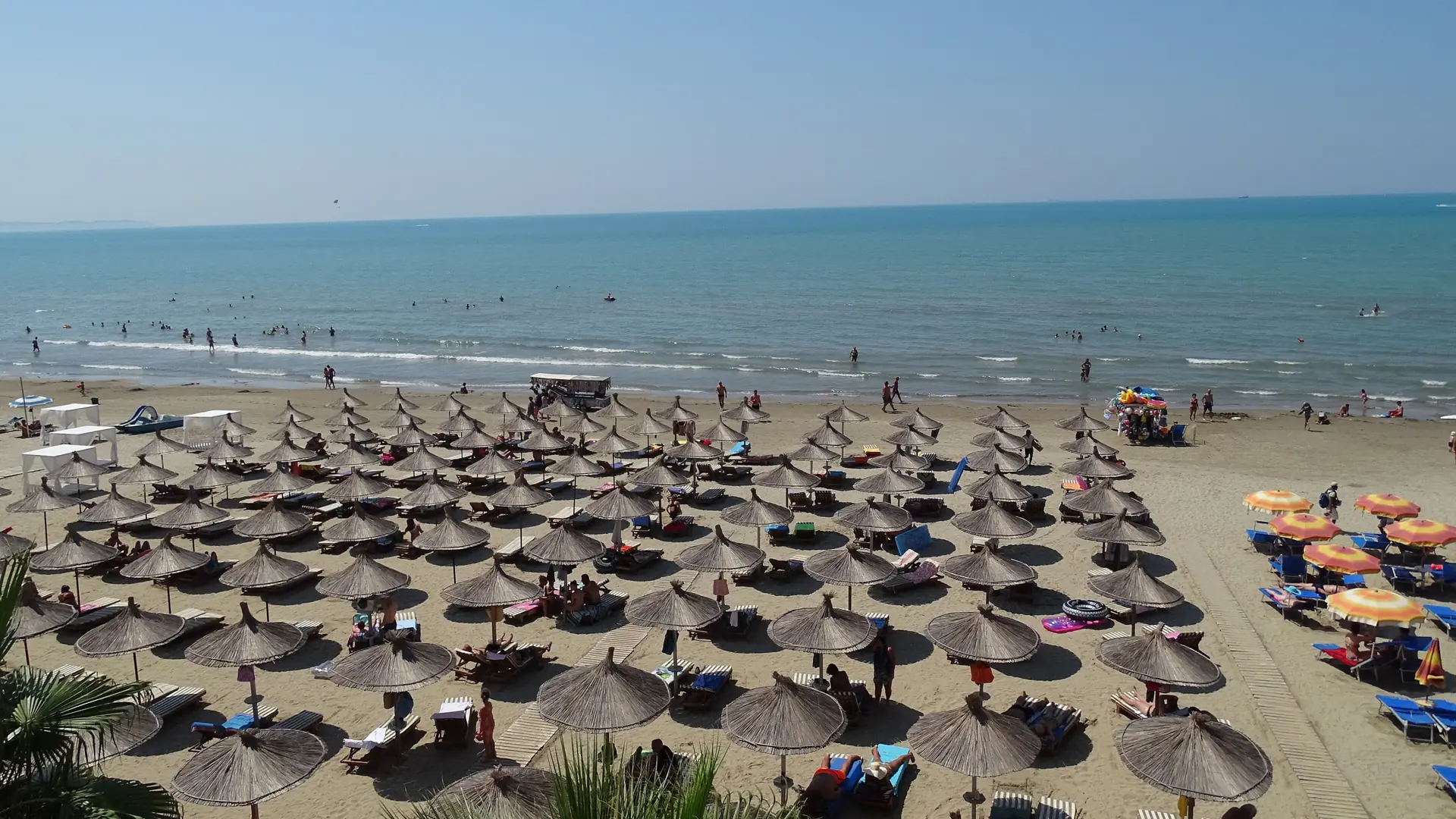Rows of thatched umbrellas on the sand at Golem, south of Durrës, beside clear shallow water