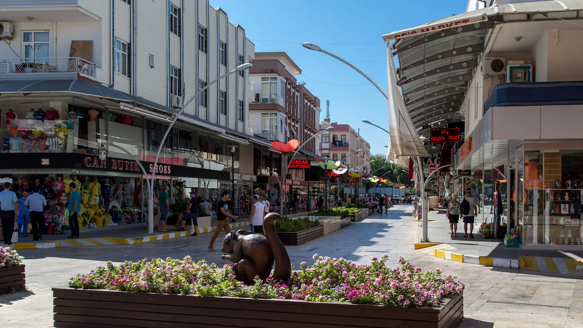 Belek town shopping street with flower planters and a bronze bear under a clear blue sky