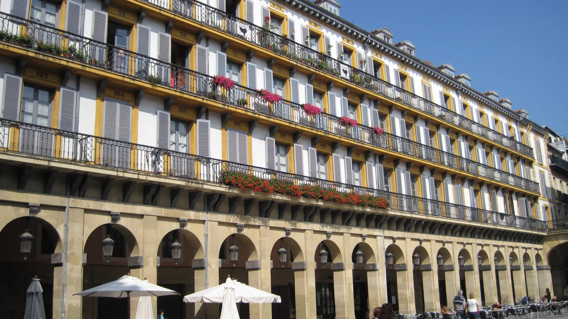 Numbered yellow balcony openings, grey shutters and stone arcades in Plaza de la Constitución