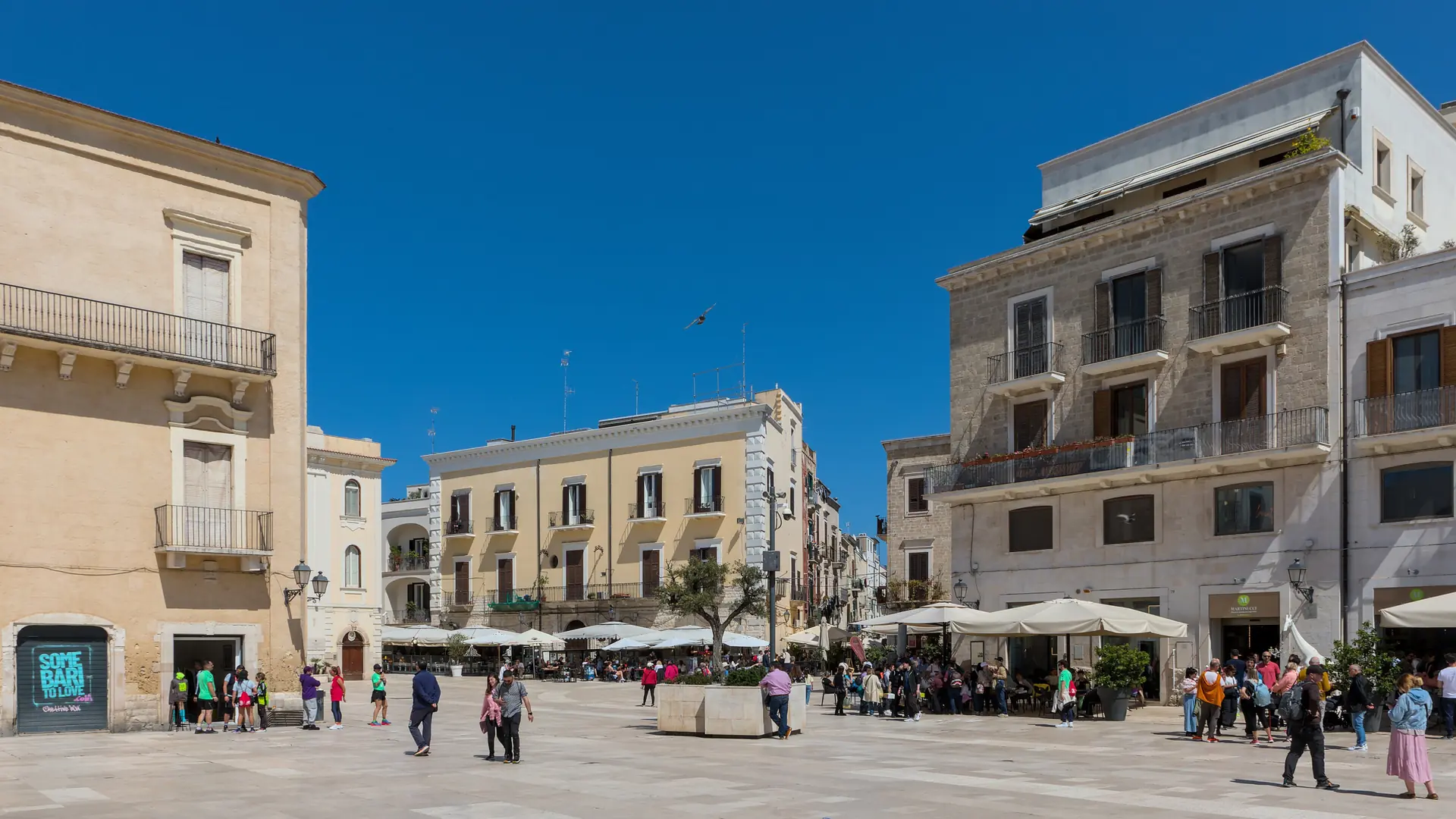 Piazza Mercantile’s pale buildings, outdoor tables and pedestrians under a blue sky in Bari.
