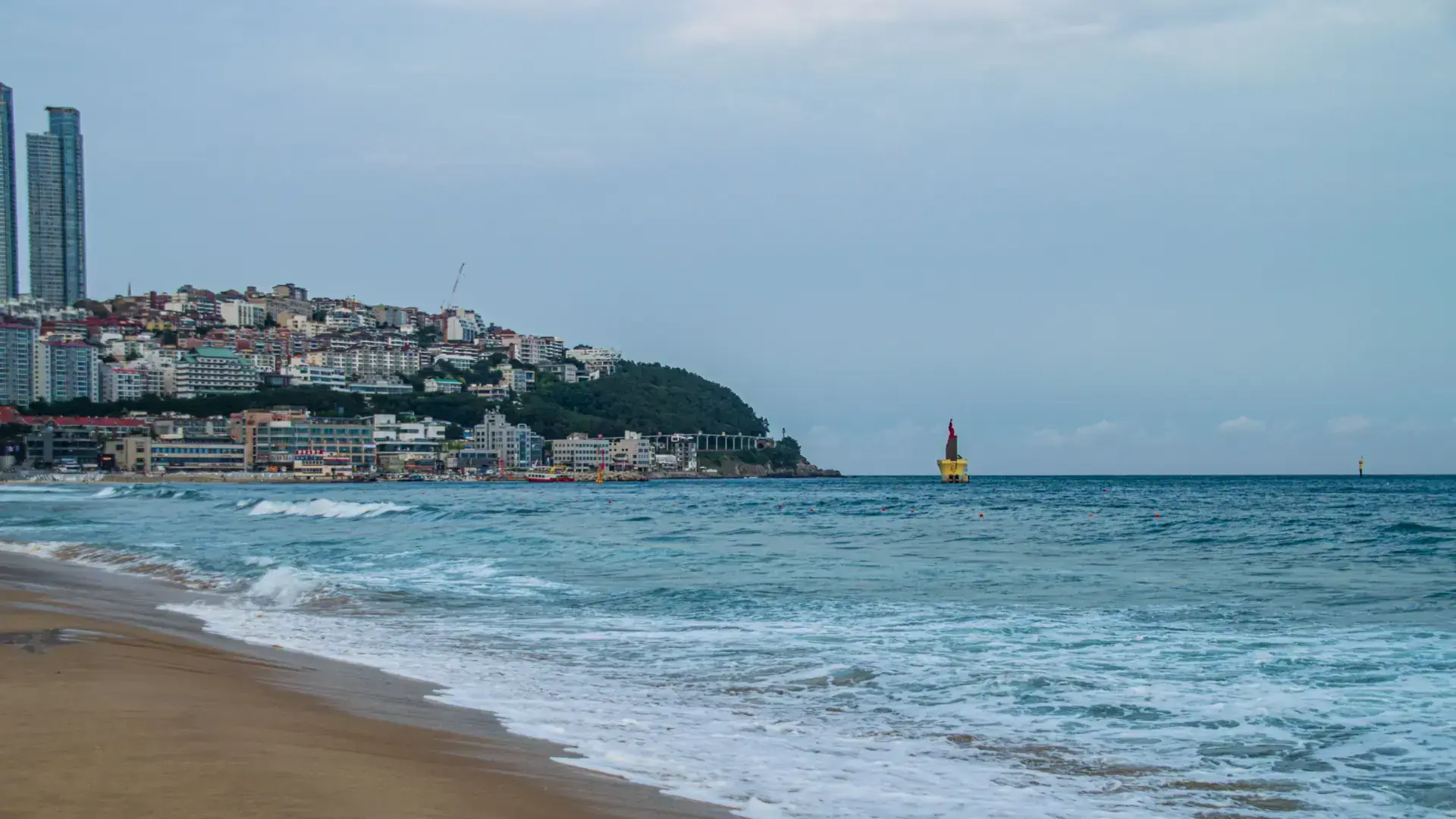 Busan coastline with a beach, waterside buildings and a yellow lighthouse beneath pale cloud