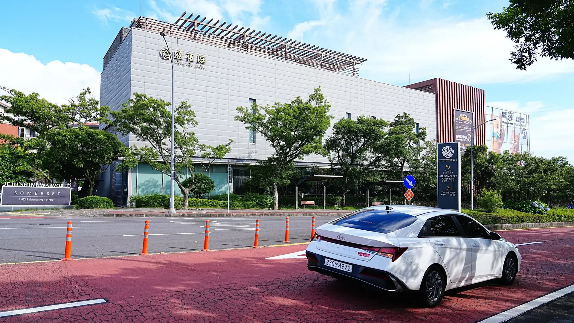 A white car and trees beside a modern shopping building on a street in Jeju City