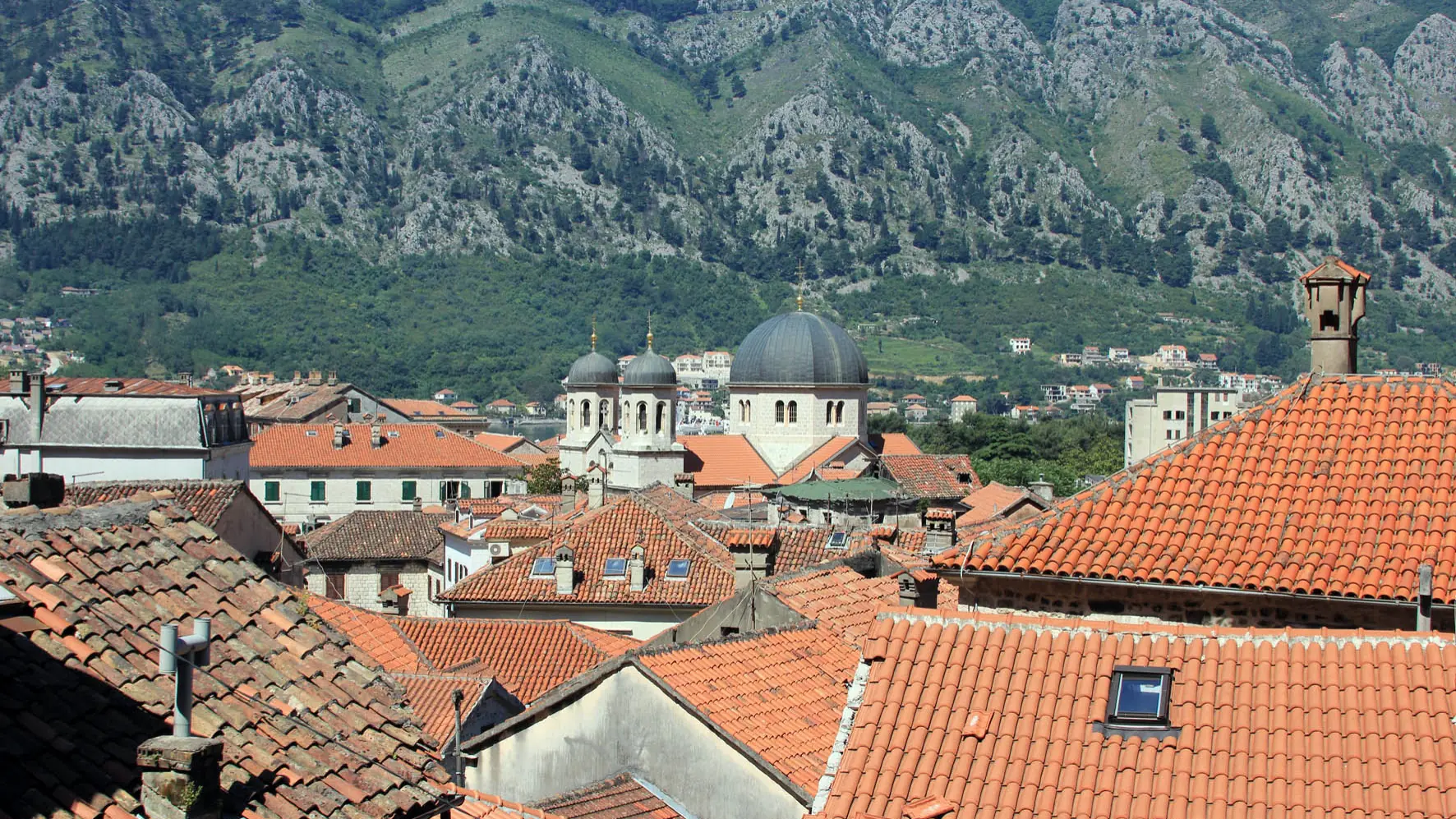Kotor Old Town rooftops and church domes beneath green mountain slopes in bright daylight