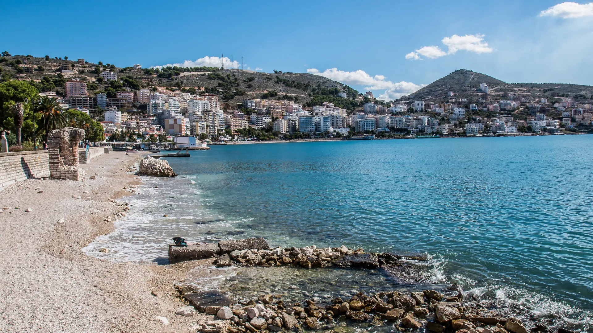 Clear turquoise water and a pebbly beach curving towards Sarandë's hillside town under a bright blue sky
