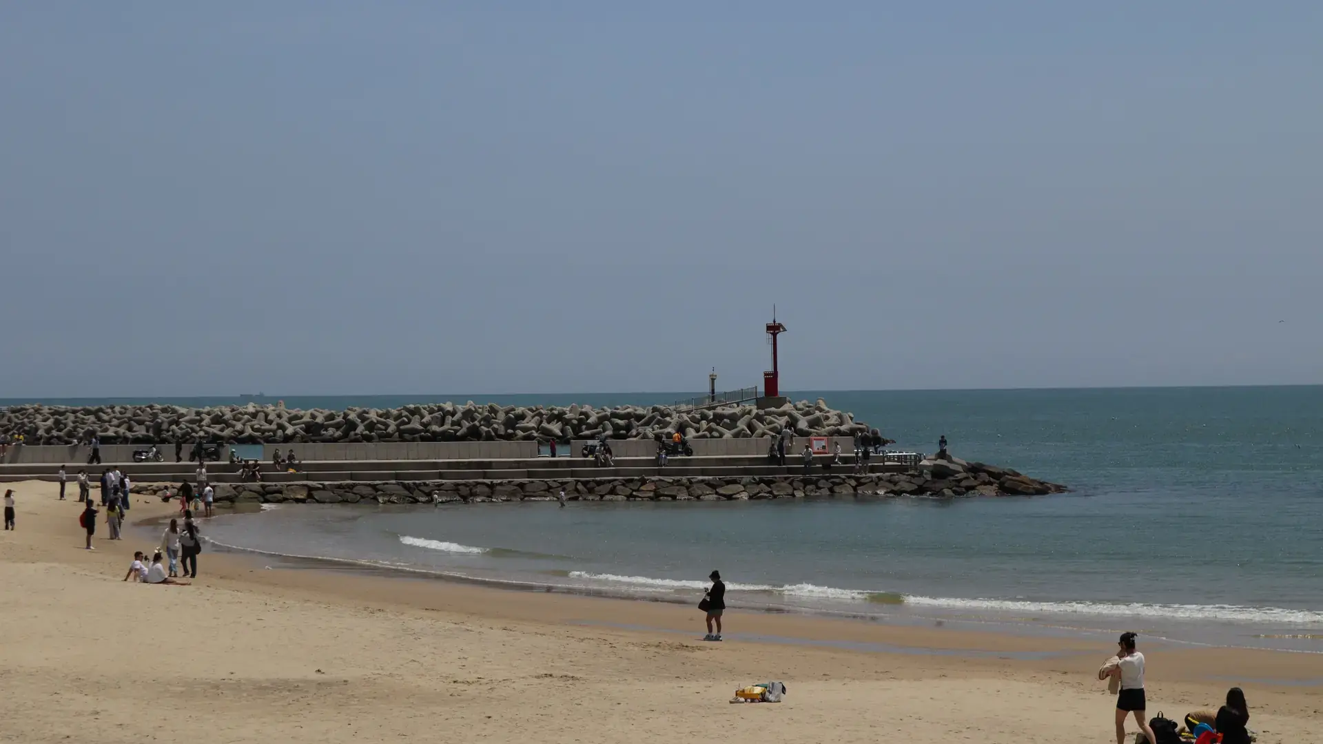 People on a sandy Busan beach beside a breakwater and red lighthouse in daylight