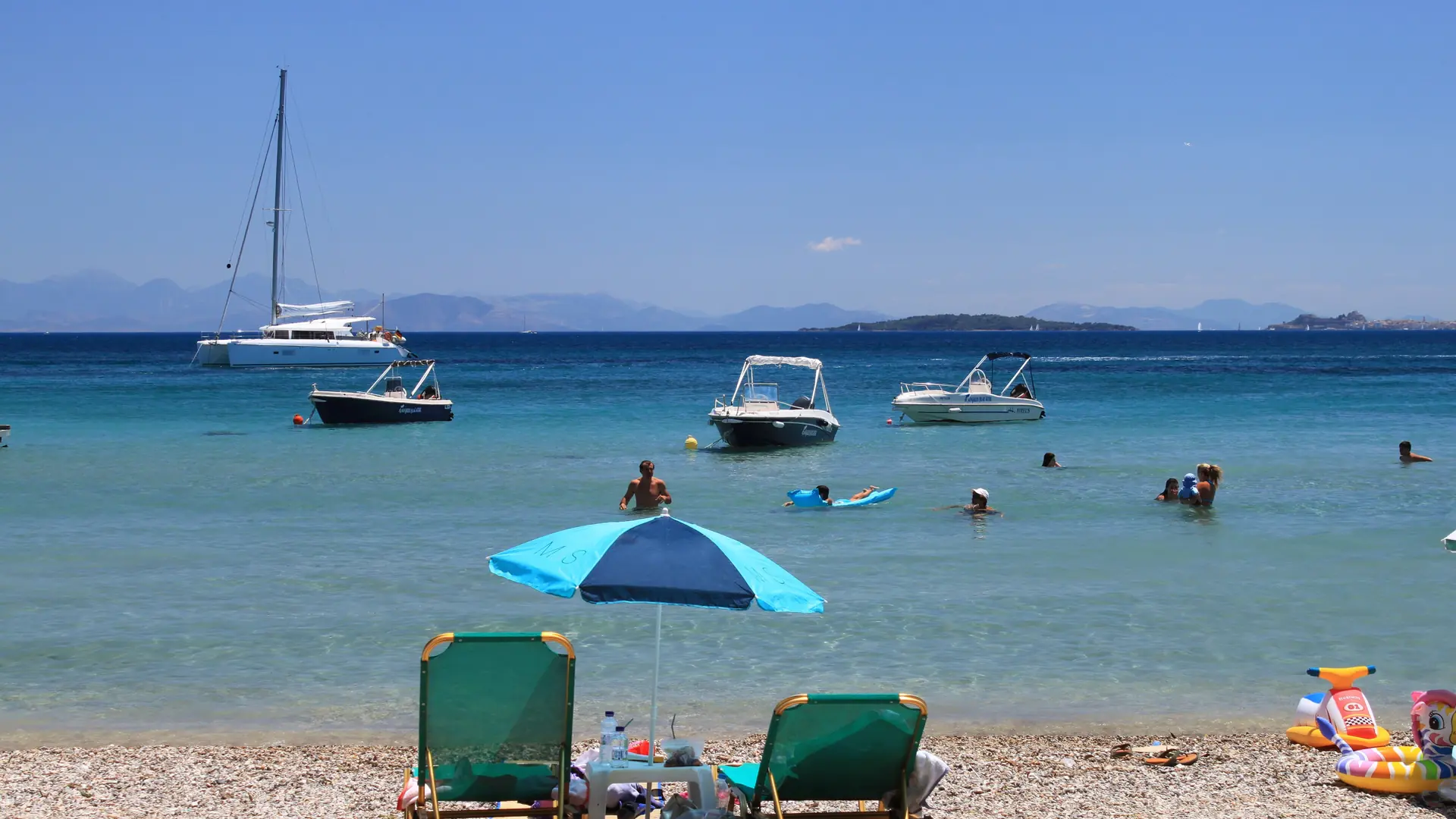 Boats and swimmers off Dassia beach, Corfu, with a blue parasol beside the water