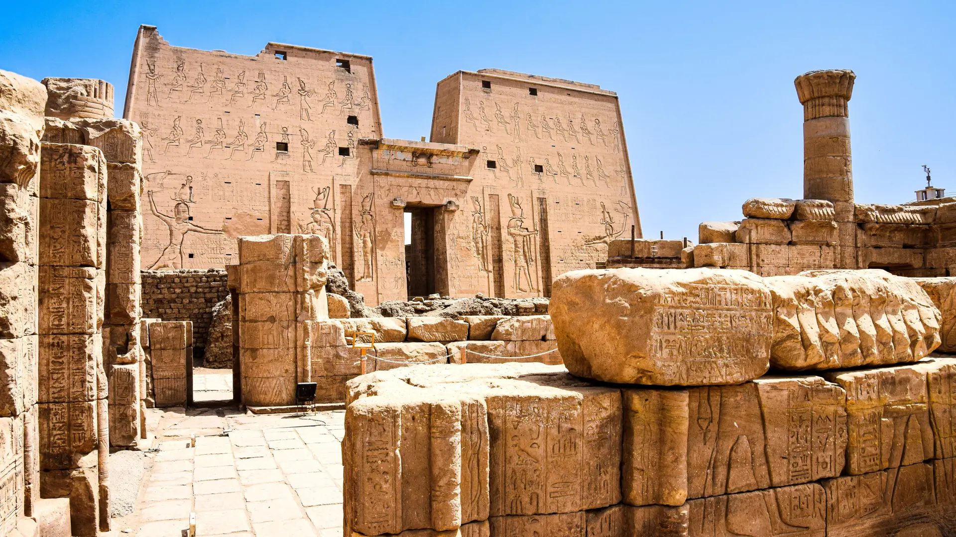 The carved pylon and stone court of Edfu Temple under a clear blue Egyptian sky.