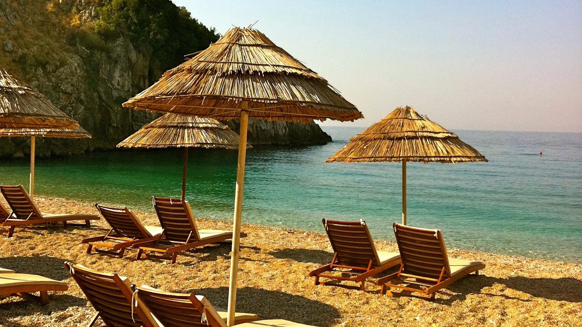 Thatched umbrellas and sunbeds on a sunny beach in Himarë beside clear turquoise water