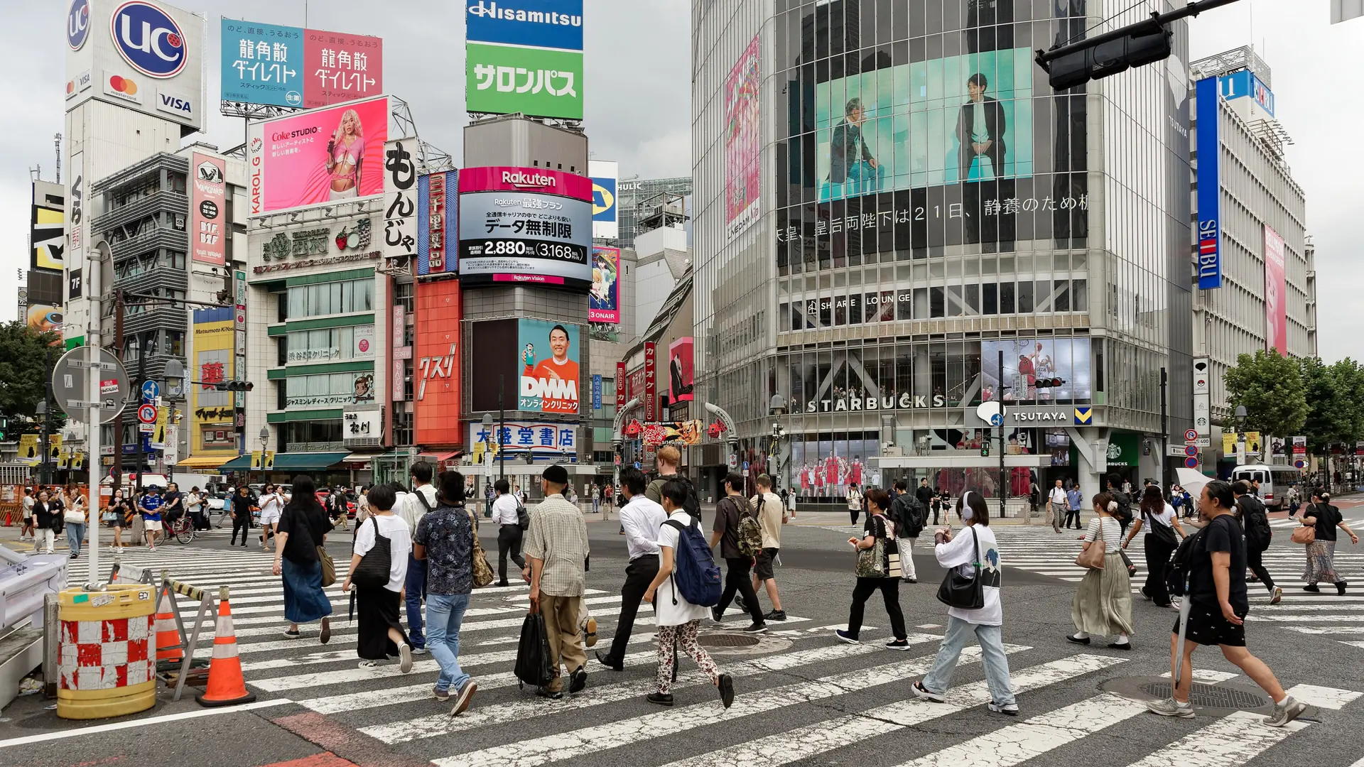 Pedestrians crossing Shibuya Scramble Crossing beside QFront’s glass facade in bright daylight.