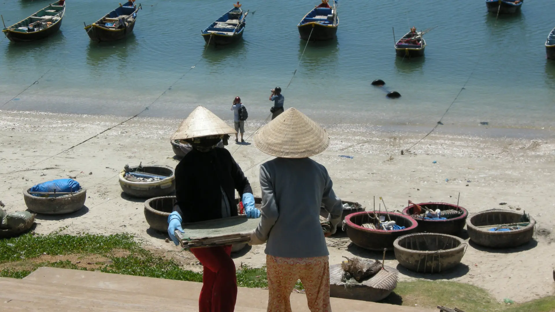 Fishing workers carrying a basket above Mui Ne harbour, a wider town scene
