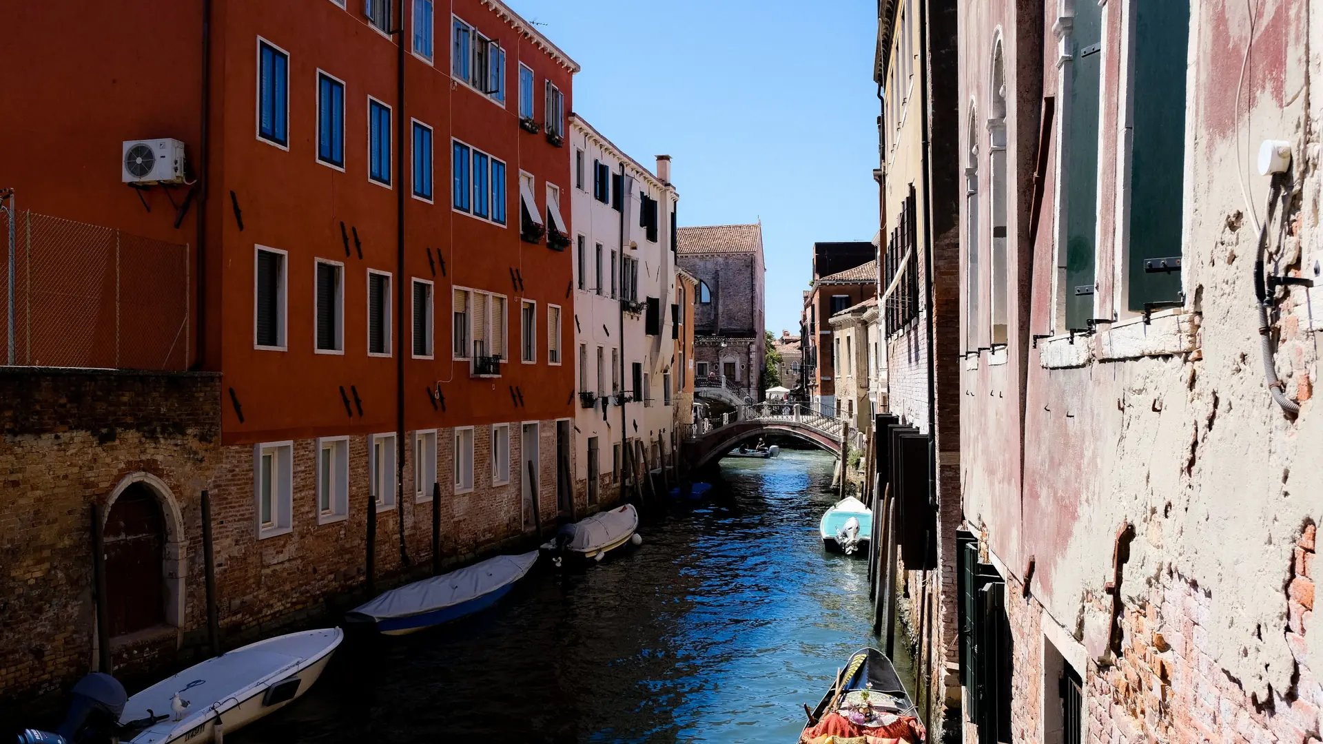 a sunlit orange-toned canal with a stone bridge and moored boats in Venice