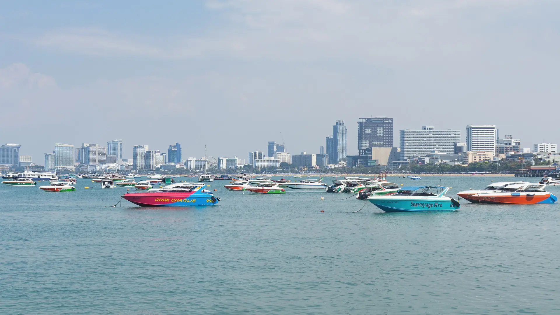 Colourful boats and the Pattaya skyline seen from the pier in daylight, rather than Naklua