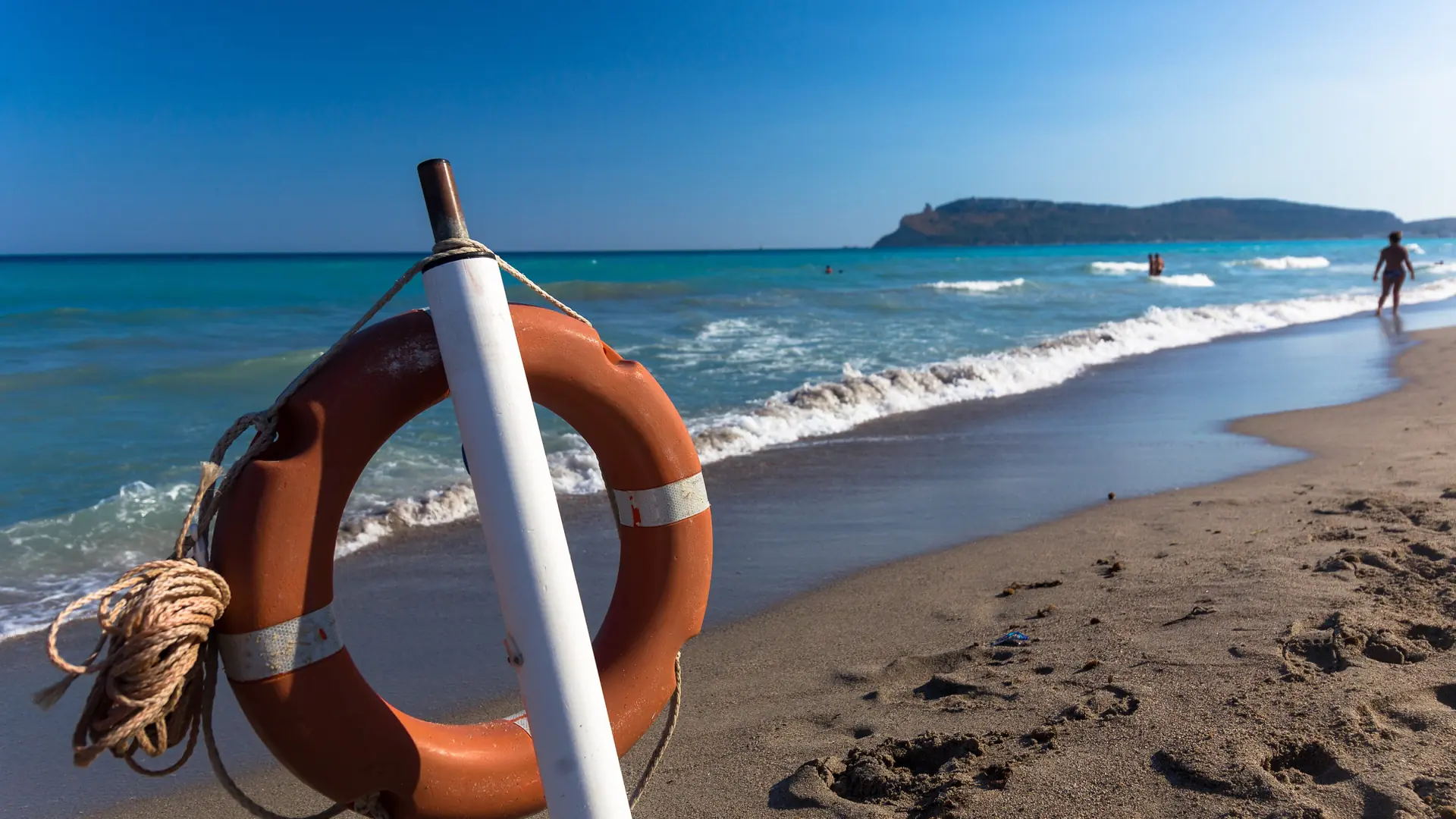 An orange lifebuoy beside Poetto’s sandy shoreline, with Cagliari’s Sella del Diavolo beyond.