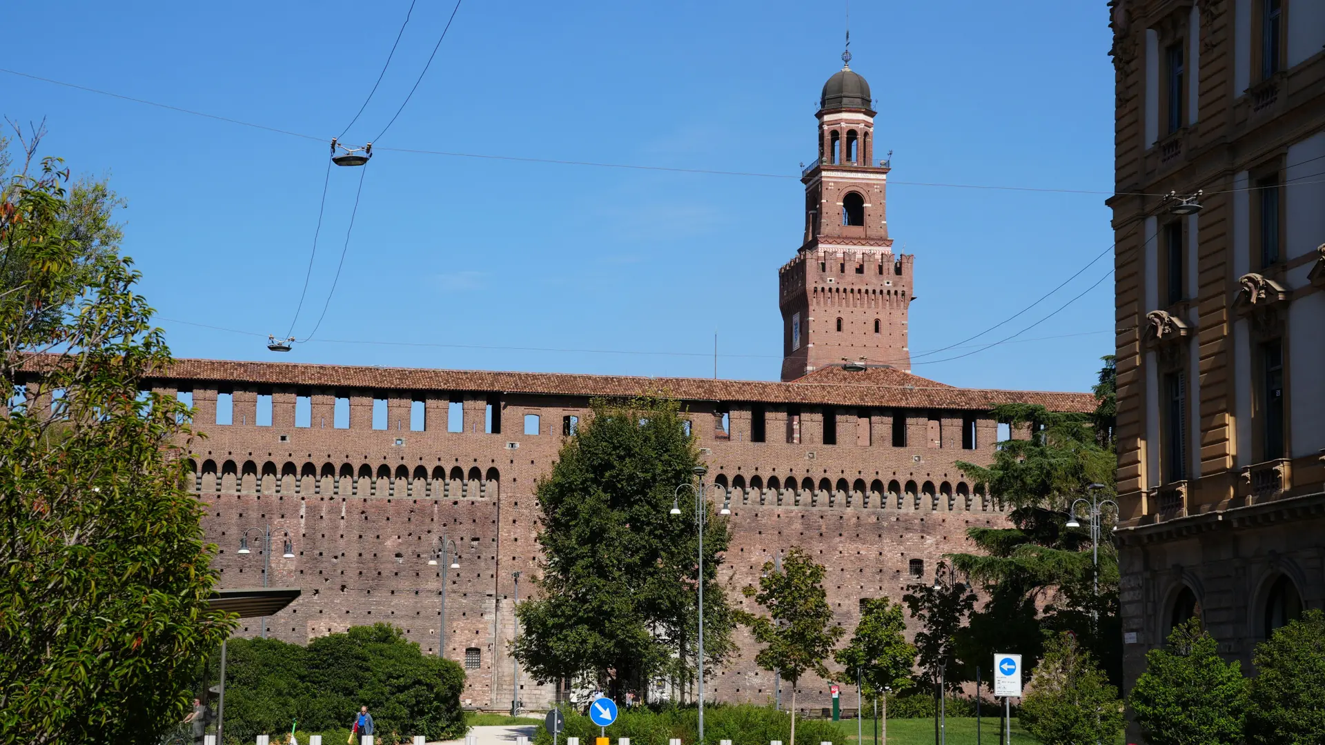 The brick walls and central tower of Castello Sforzesco in Milan beneath a blue sky.
