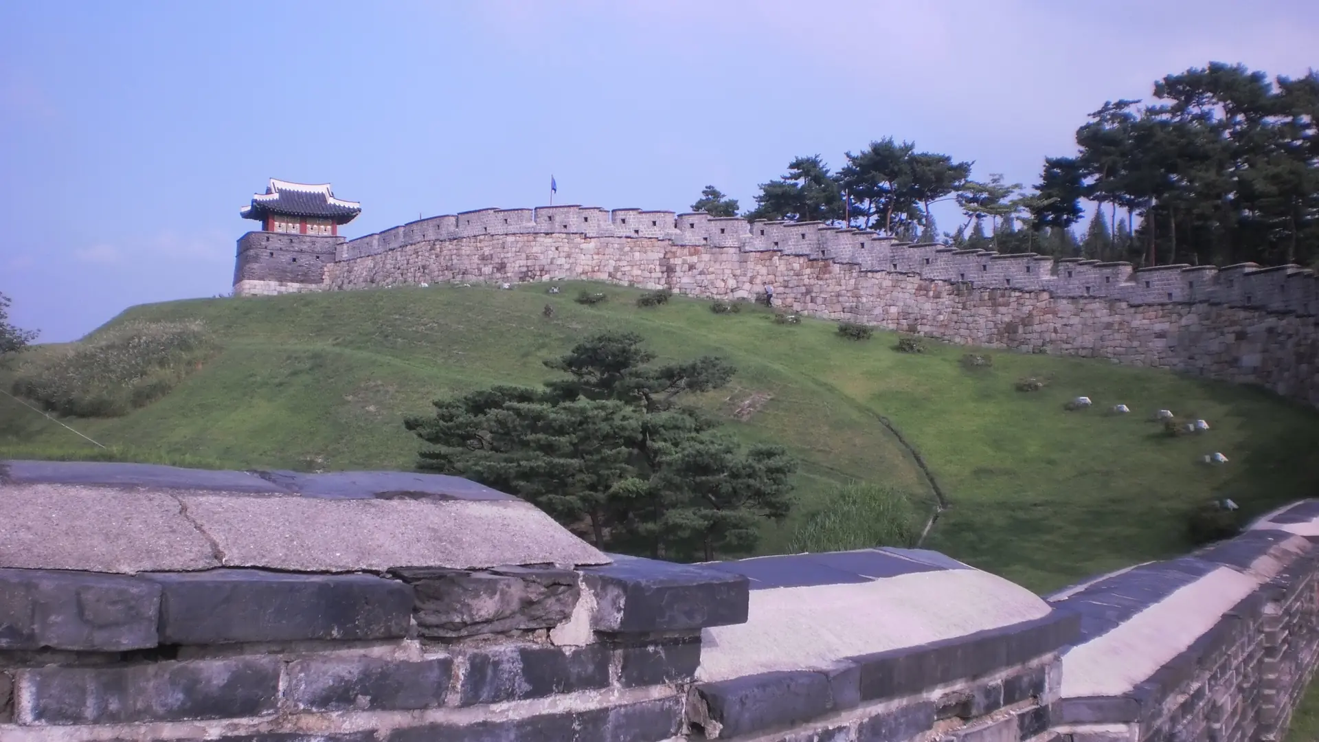 Hwaseong Fortress's stone wall and a red-and-white watchtower pavilion atop a grassy hill under a clear blue sky, Suwon