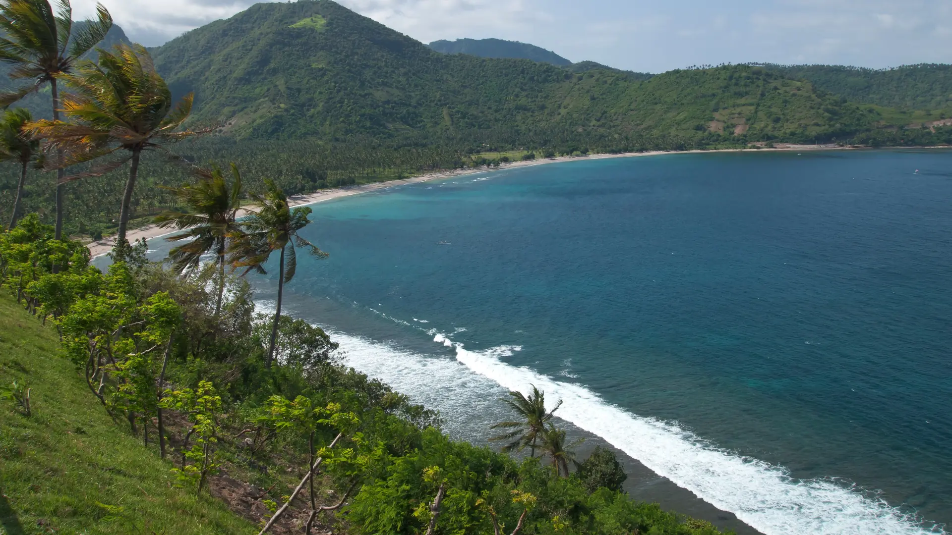 Wider Lombok coastal view of a curving beach, turquoise water and palm-covered green hills