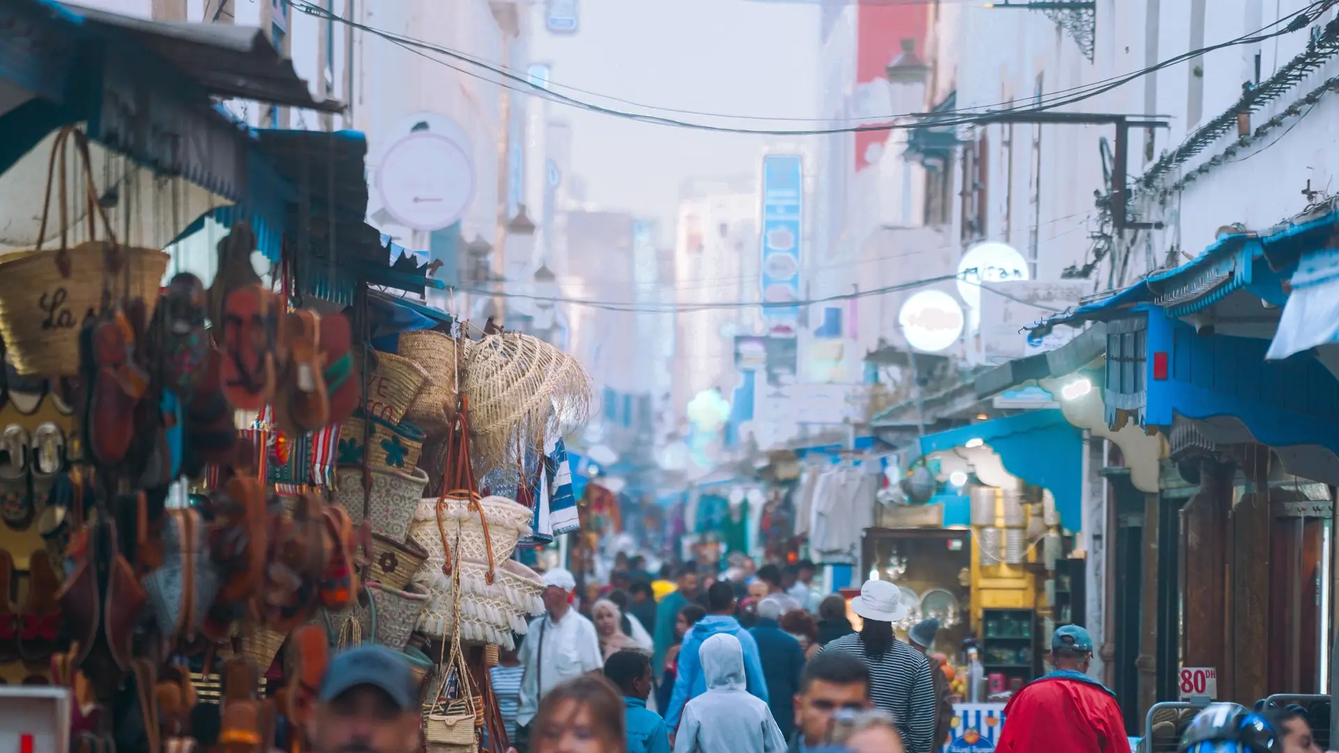 Shoppers and stalls along a busy Essaouira medina market lane in daylight