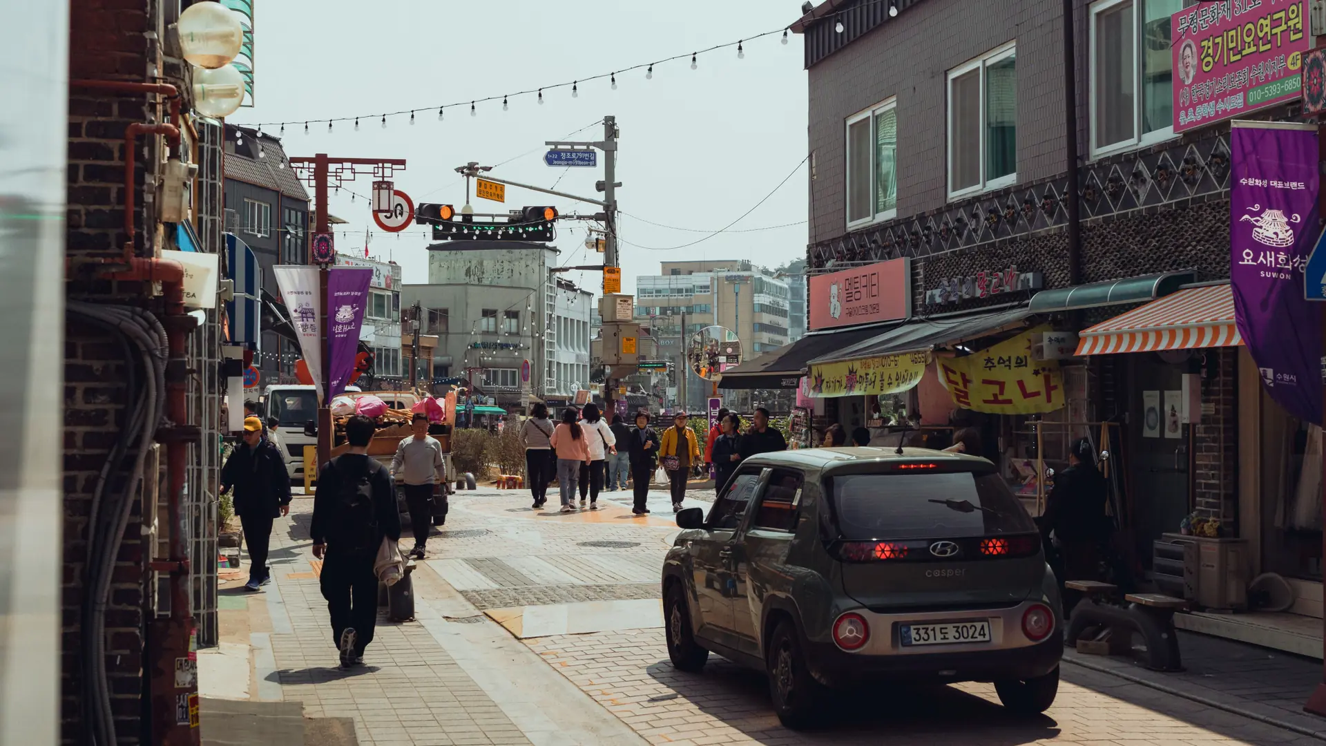 A Suwon shopping street with pedestrians, shop signs and overhead banners in bright daylight; city context