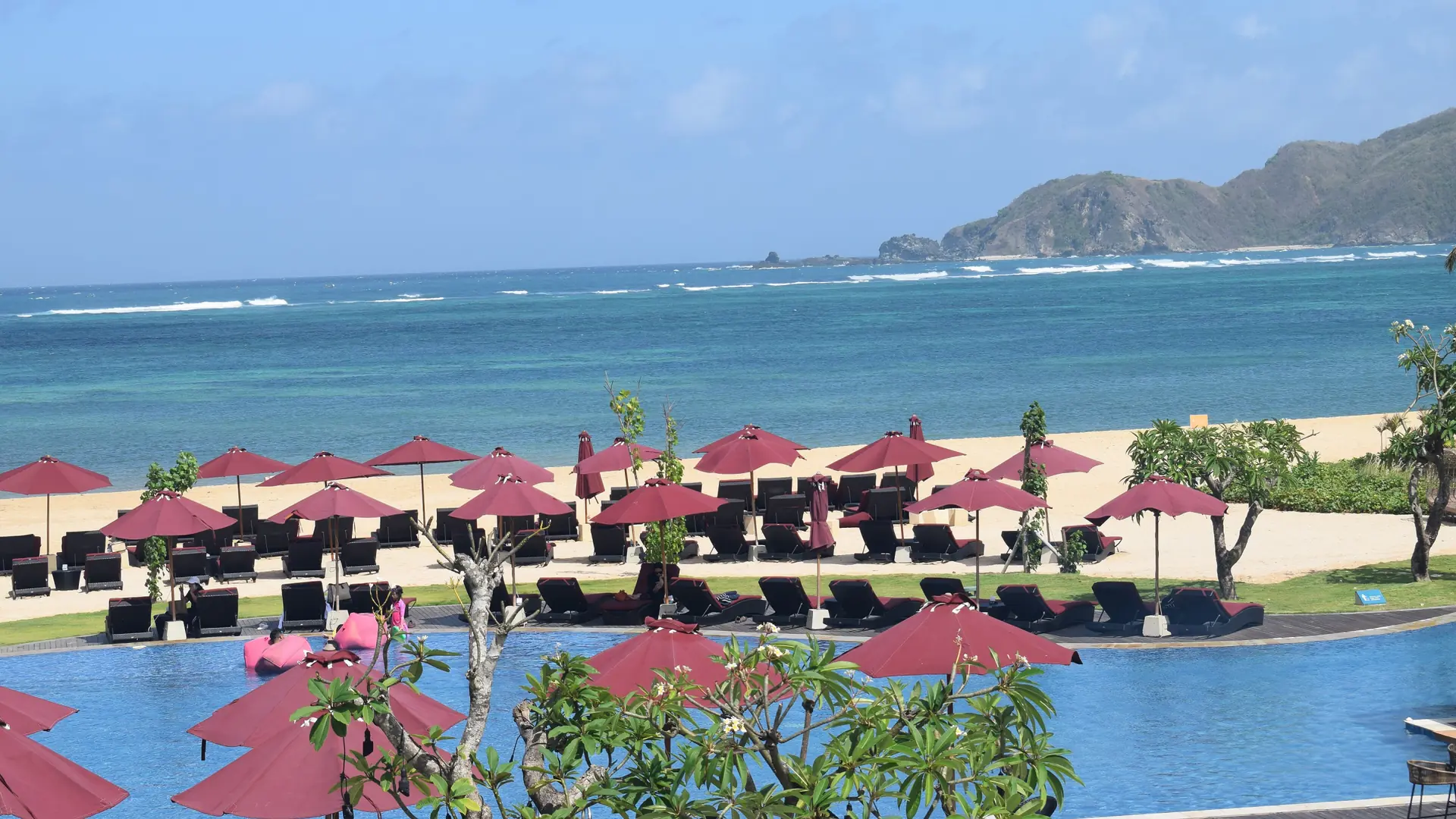 Pool, red beach umbrellas and Mandalika’s blue water seen from Pullman Lombok Mandalika Beach Resort