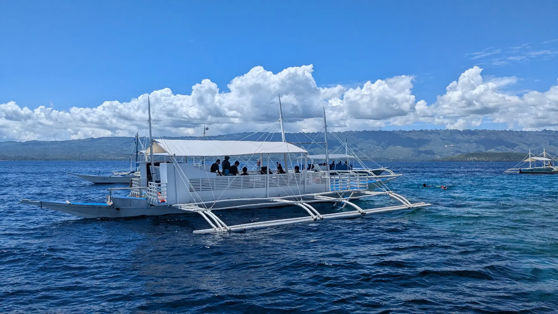 A white outrigger boat and swimmers on blue water off Moalboal, with a mountainous coast beyond
