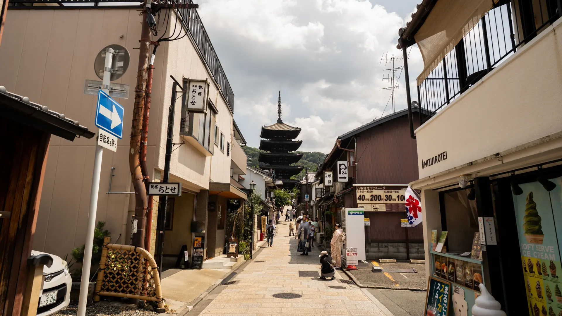 A sunlit Kyoto street leading towards Yasaka Pagoda, with shops and visitors