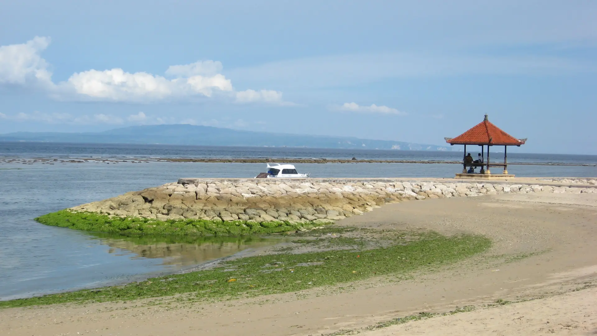 A red-roofed seaside pavilion and exposed shoreline at low tide in Sanur