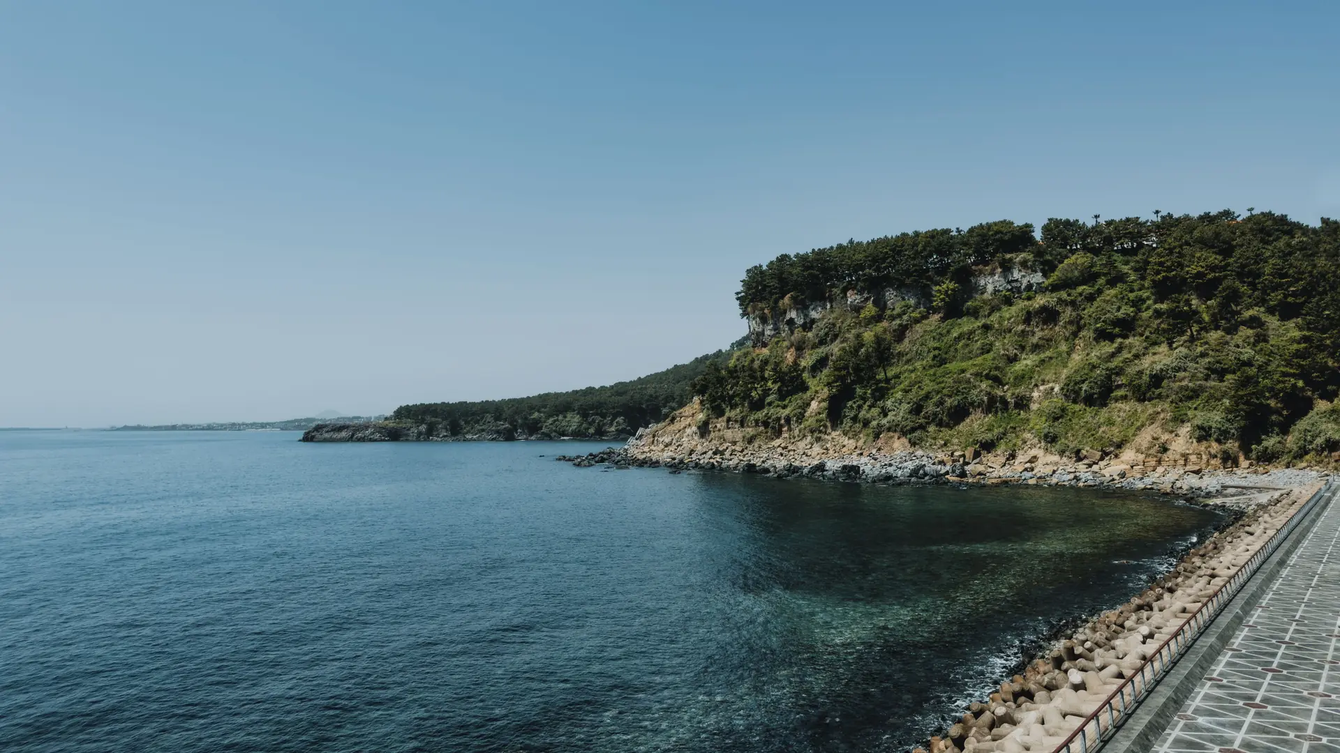 Paved waterfront path beside a blue bay and wooded headland on Jeju Island