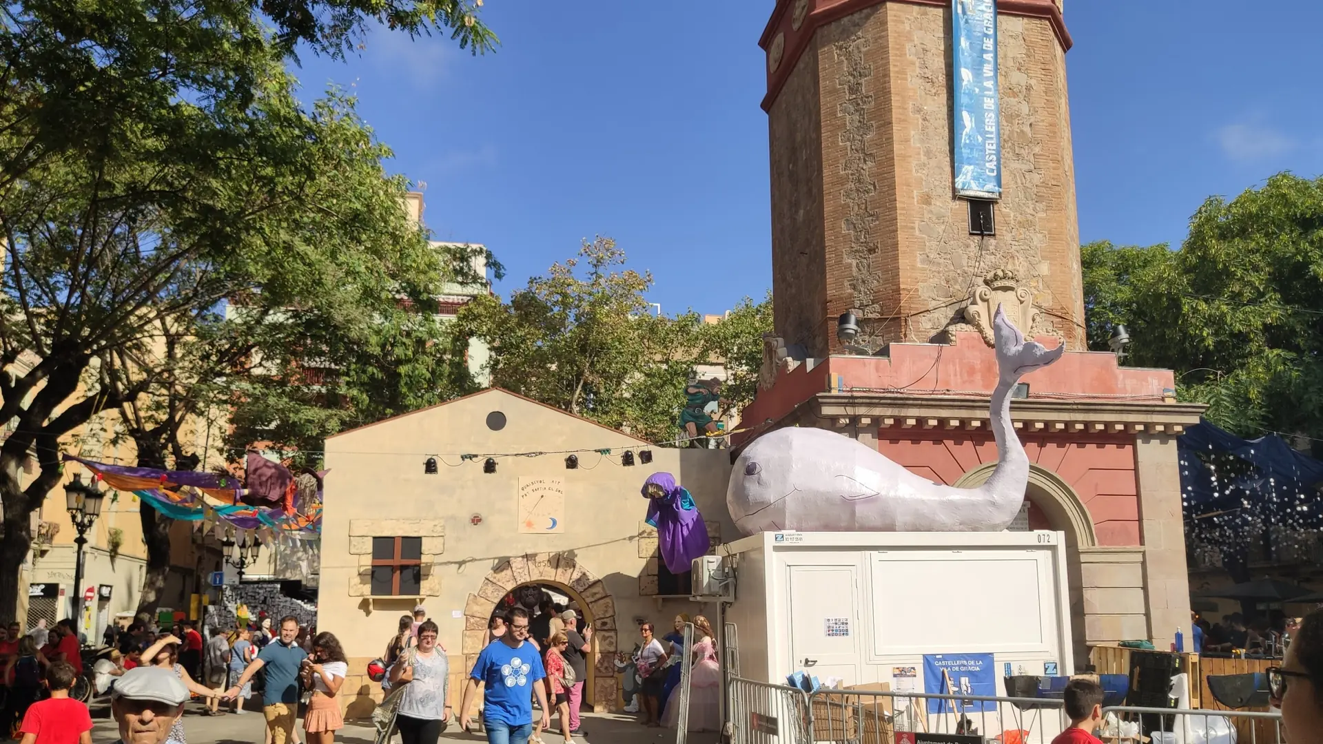 Visitors and festival decorations beside the clock tower on Plaça de la Vila de Gràcia during the 2023 Festa Major